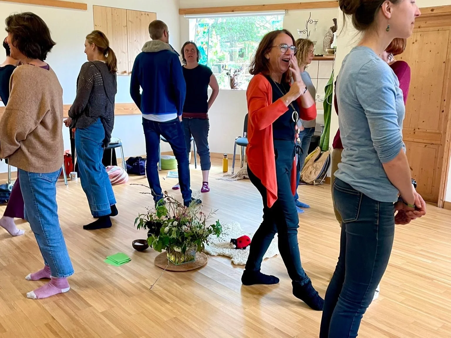 A group of people standing in a circle indoors, some smiling and laughing, participating in a group activity or meditation. The room has wooden walls and flooring, with a window letting in natural light. There are casually placed personal items and plants on the floor.
