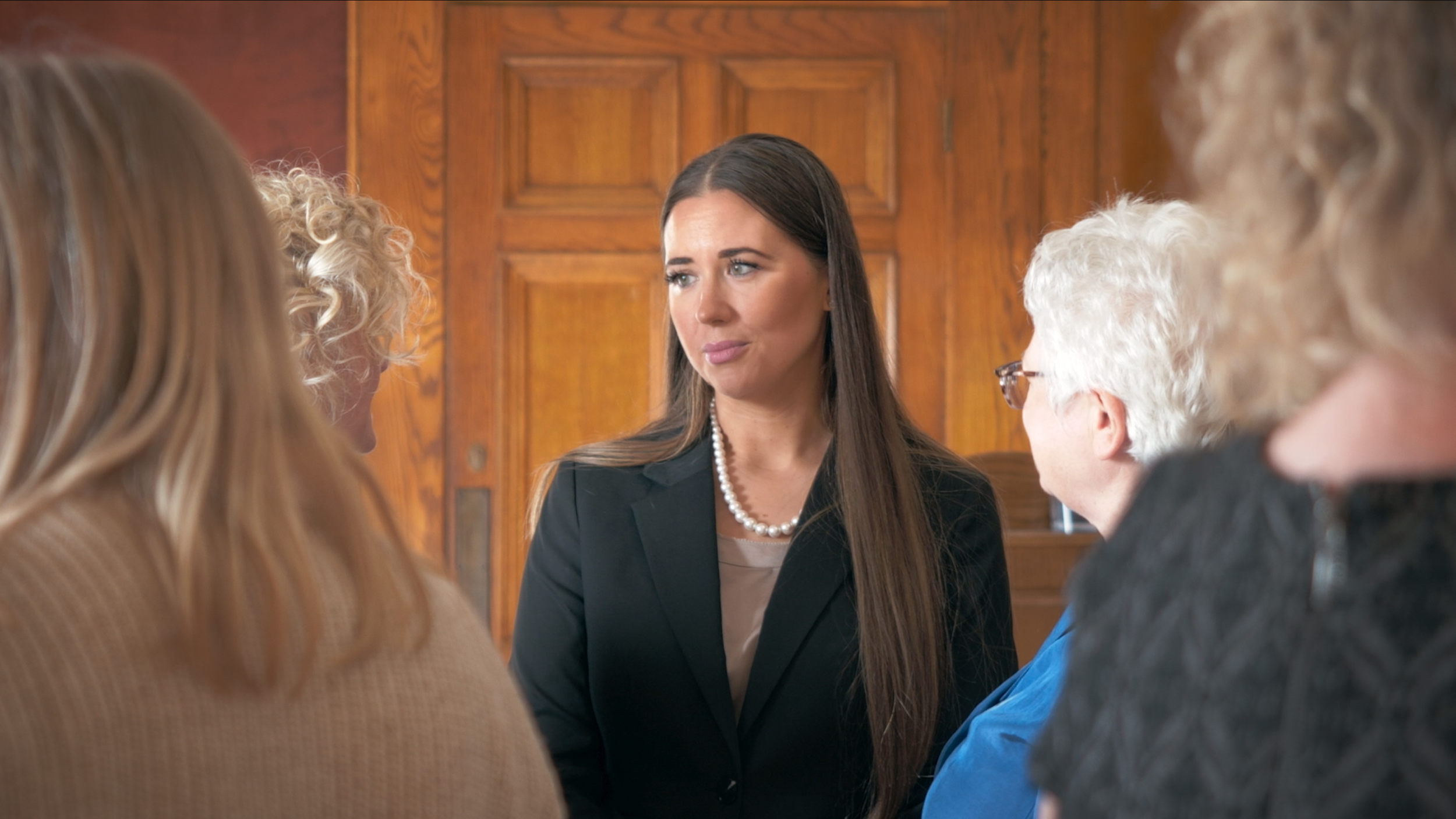 Woman with long brown hair and wearing a pearl necklace in conversation with elderly women in a room with wood-paneled walls.
