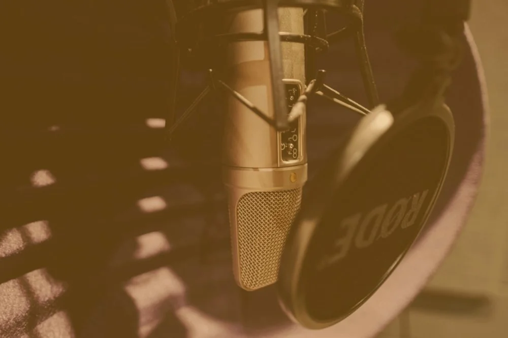 A close-up of a vintage microphone and a Rode headphone resting on a surface, with shadows of a lamp on the surface.