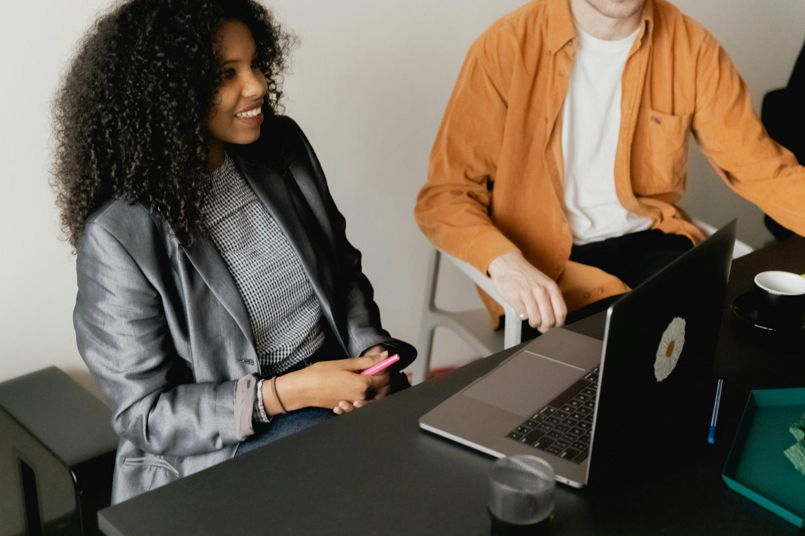 Two colleagues talking at a desk with a laptop, one smiling in a gray blazer.