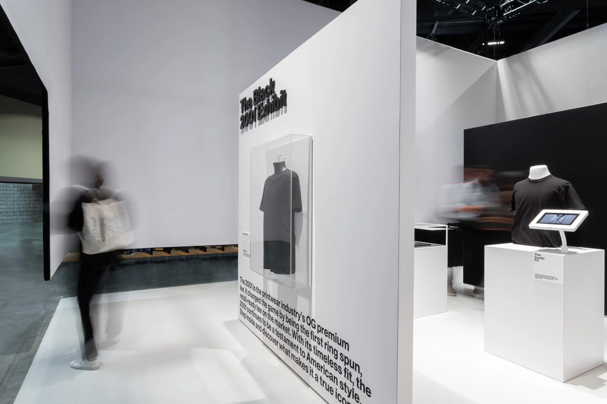 Exhibit at a retail or museum space with a sign that reads 'The Black 2001 Tee' and displayed clothing items. Blurred visitors walk past, and the display includes a description about the 2001 tee and its significance in the fashion industry.
