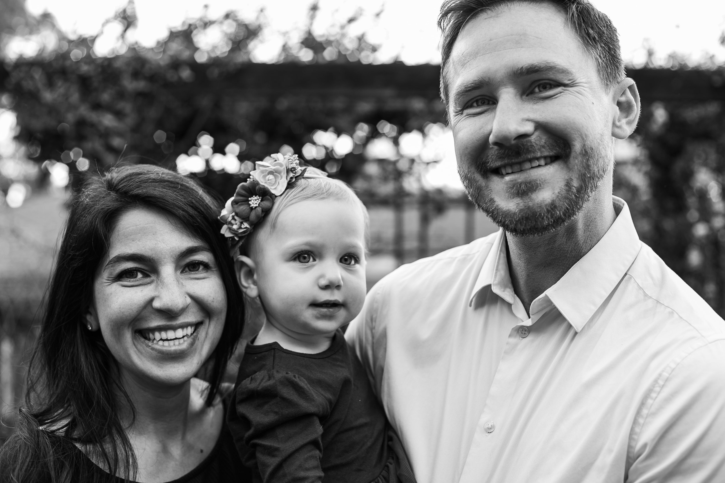 Black-and-white photo of a happy family consisting of a woman, a man, and a young girl. The woman has long hair, and the girl is wearing a floral headband. The family is smiling outdoors with trees in the background.