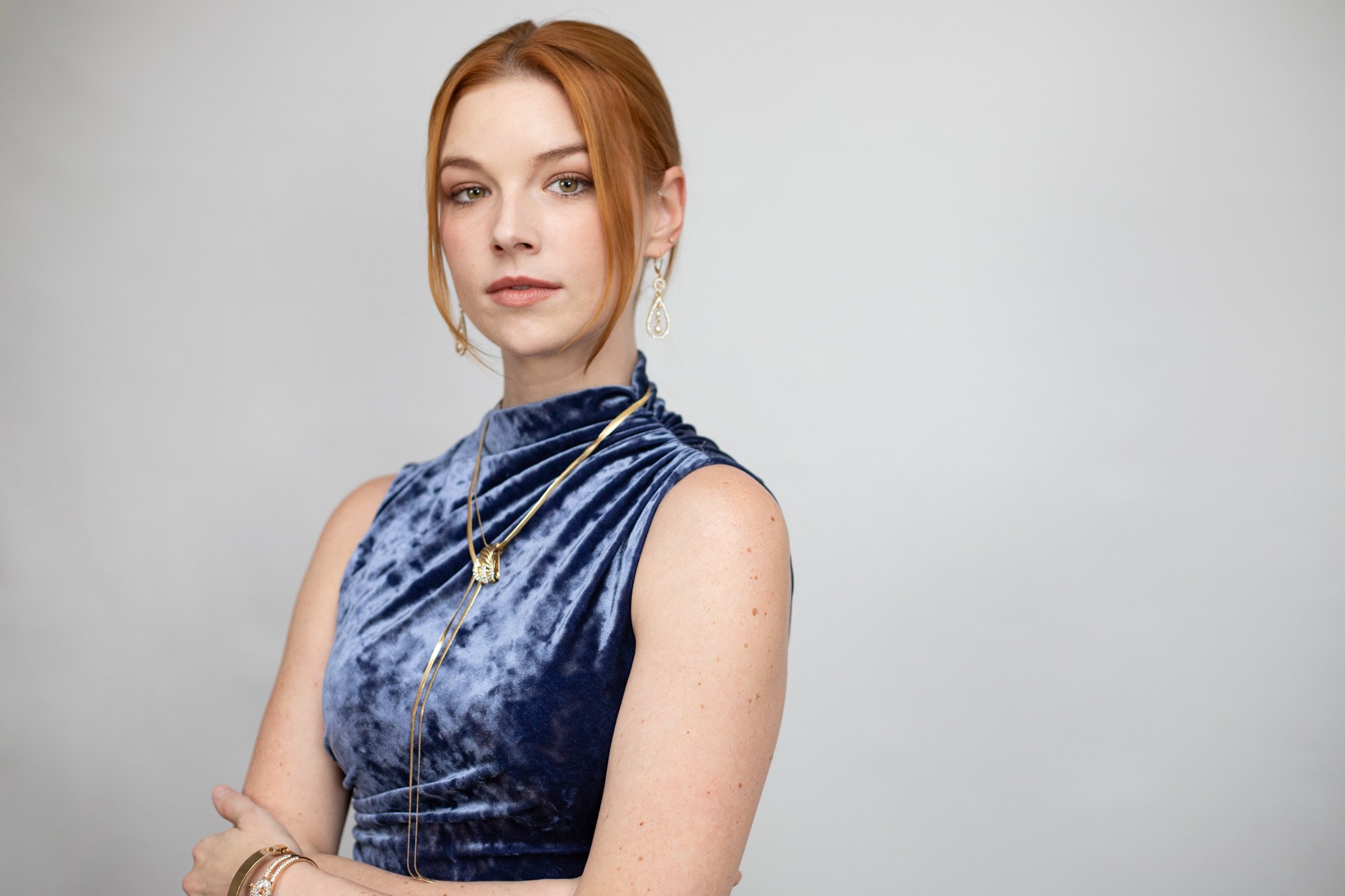 Red-haired woman in a velvet blue dress with jewelry against a plain light gray background.