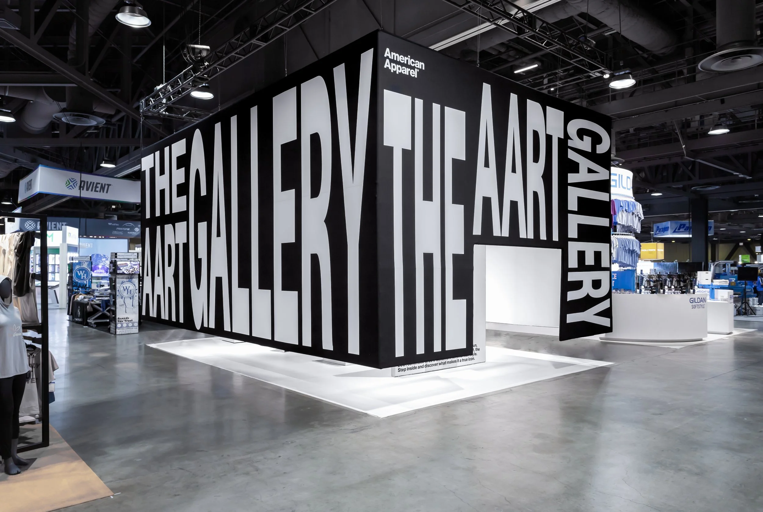 Large black and white display with the word 'THE GALLERY OF APPAREL' in bold, stylized letters at an indoor trade show or exhibition. The background shows various booths and textile products.