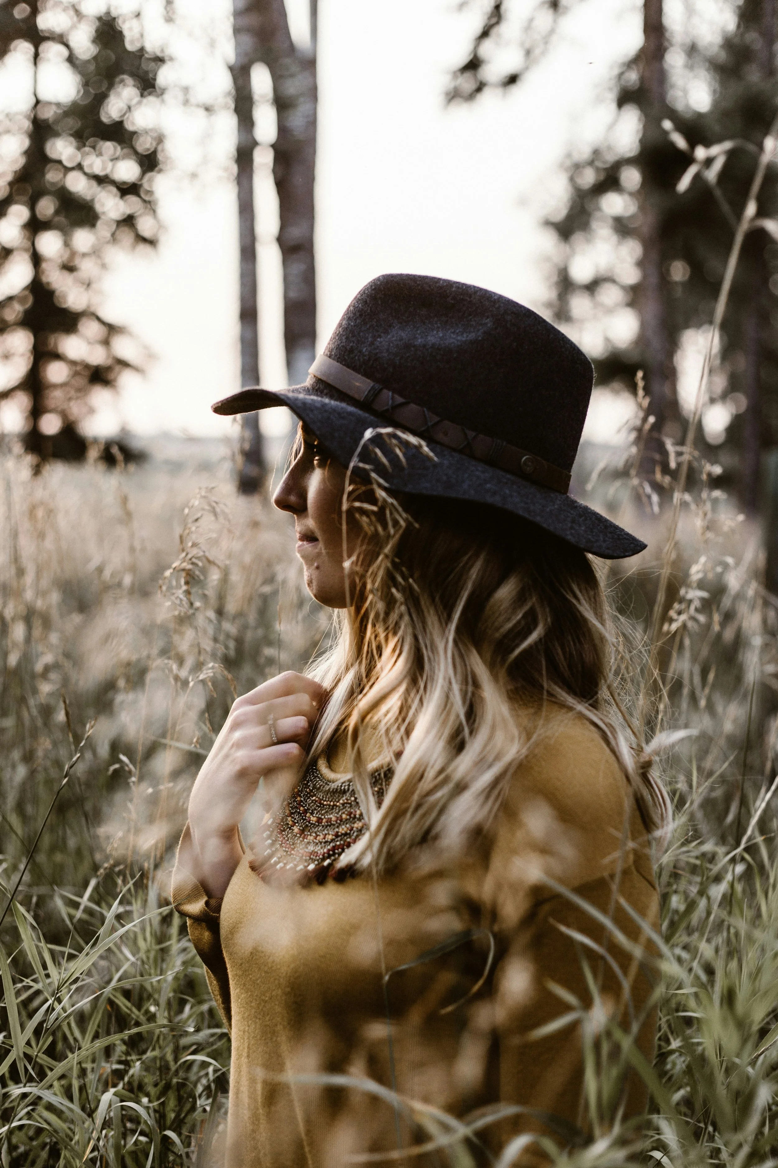A woman in a brown coat and black wide-brimmed hat standing in a field of tall grass with trees in the background at sunset.