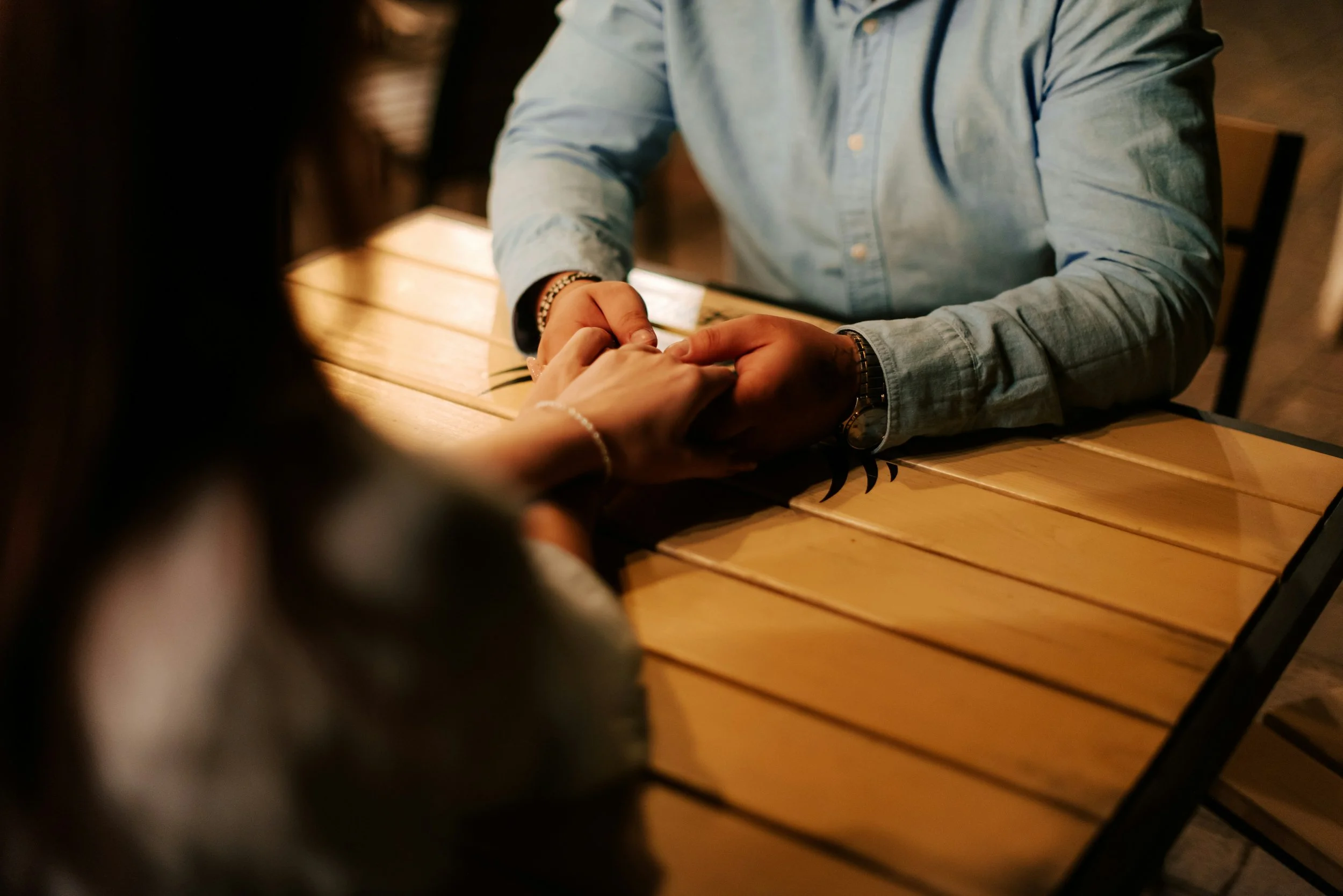A man in a light blue shirt holding hands with a woman at a wooden table in a cozy, dimly lit setting.