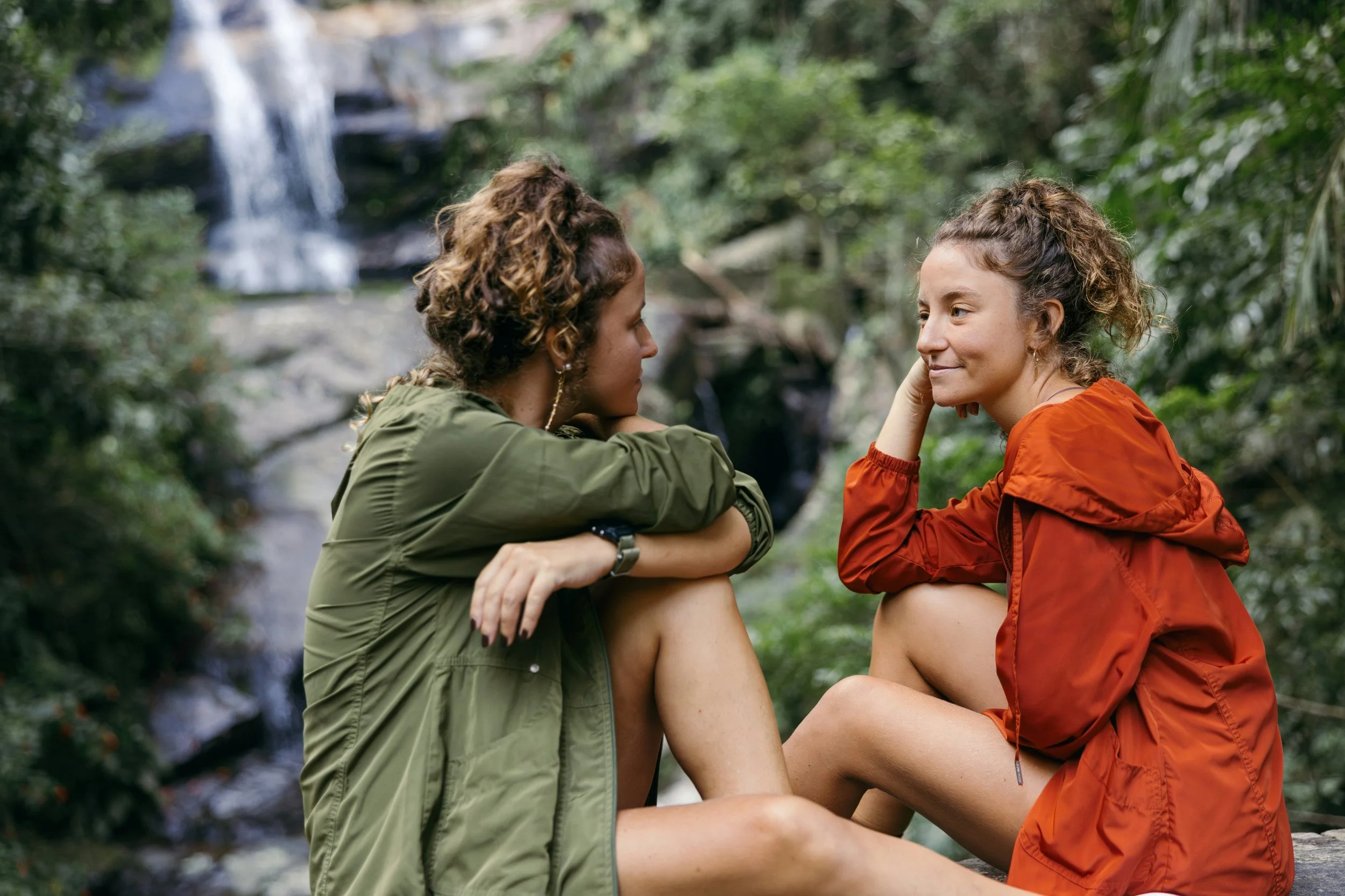 Two women sitting outdoors in a forest, facing each other and smiling, with a waterfall in the background.