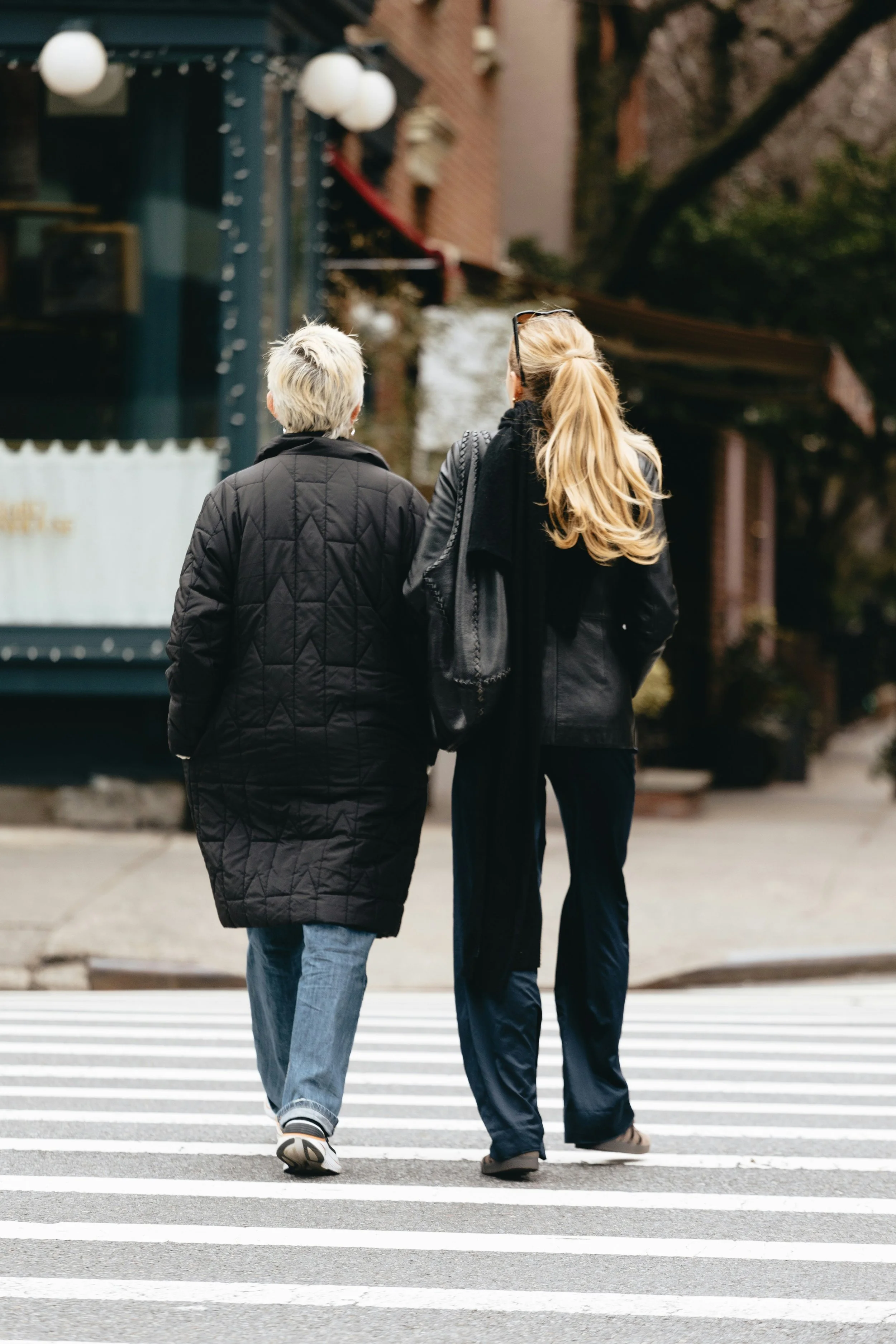 Two women walking across a crosswalk in an urban neighborhood, one with short gray hair wearing a black quilted coat and jeans, the other with long blonde hair in a ponytail wearing a black jacket and black pants.