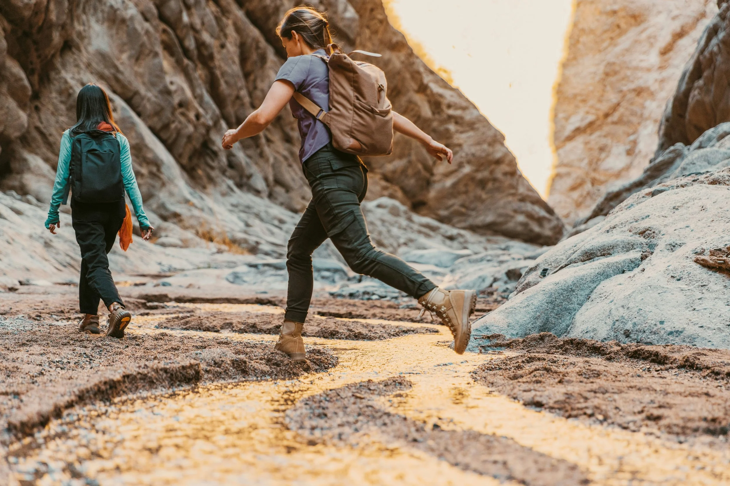 Two women hiking through a rocky canyon, wearing backpacks, with high-energy terrain and sunlight filtering through the canyon walls.