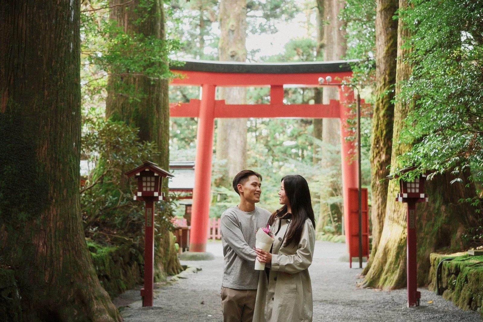 A couple standing in a forest with a large red gate behind them, one person holding a bouquet, smiling at each other.