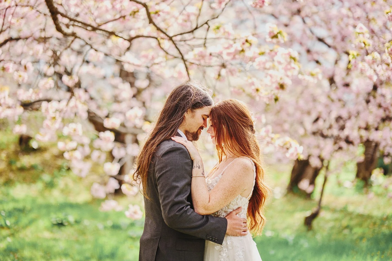 A couple with long hair and dressed in wedding attire embrace outdoors among blooming cherry blossom trees.