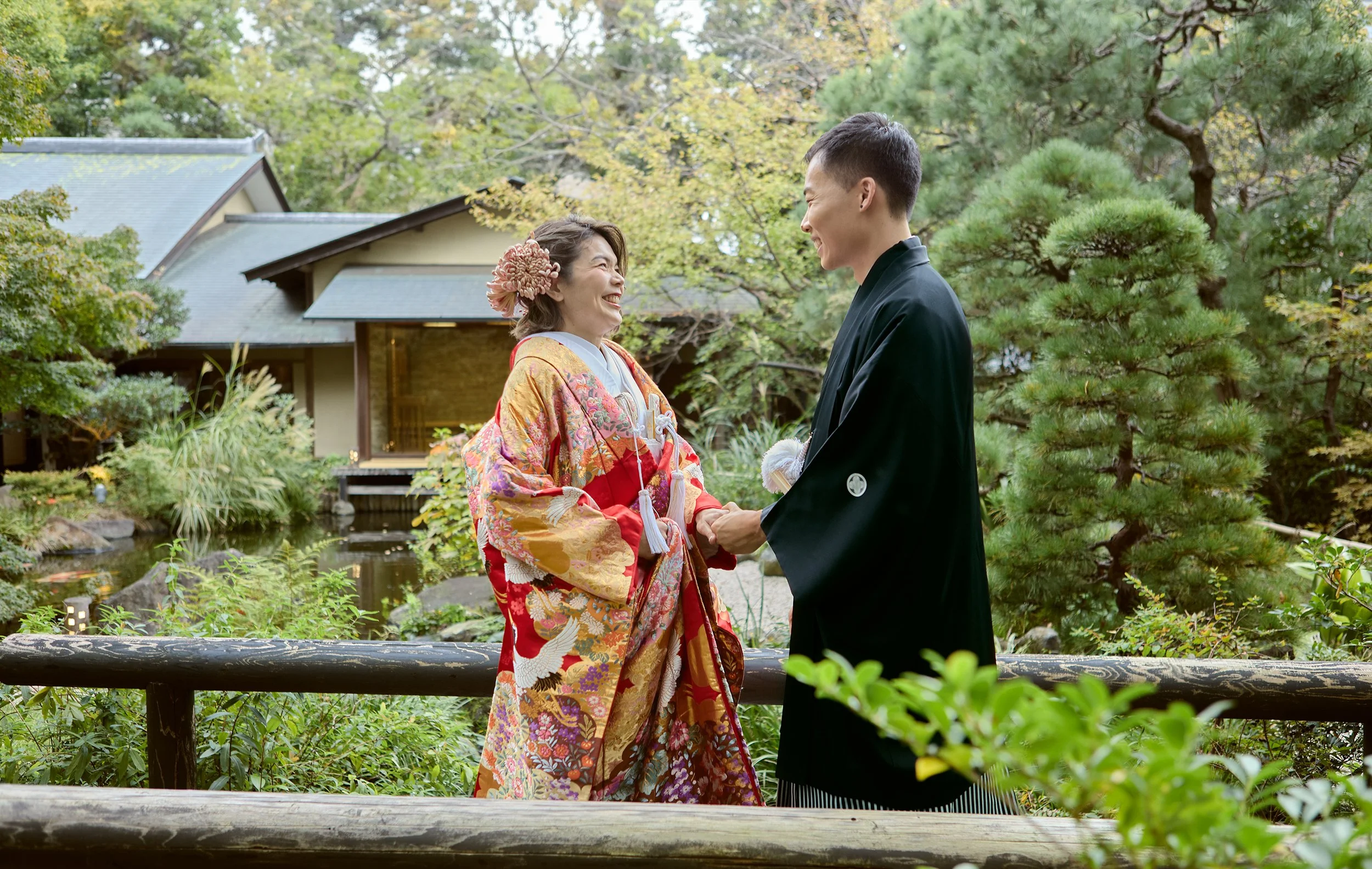 A couple in traditional Japanese wedding attire standing on a bridge in a garden, smiling and holding hands, with a pond and traditional Japanese buildings in the background.