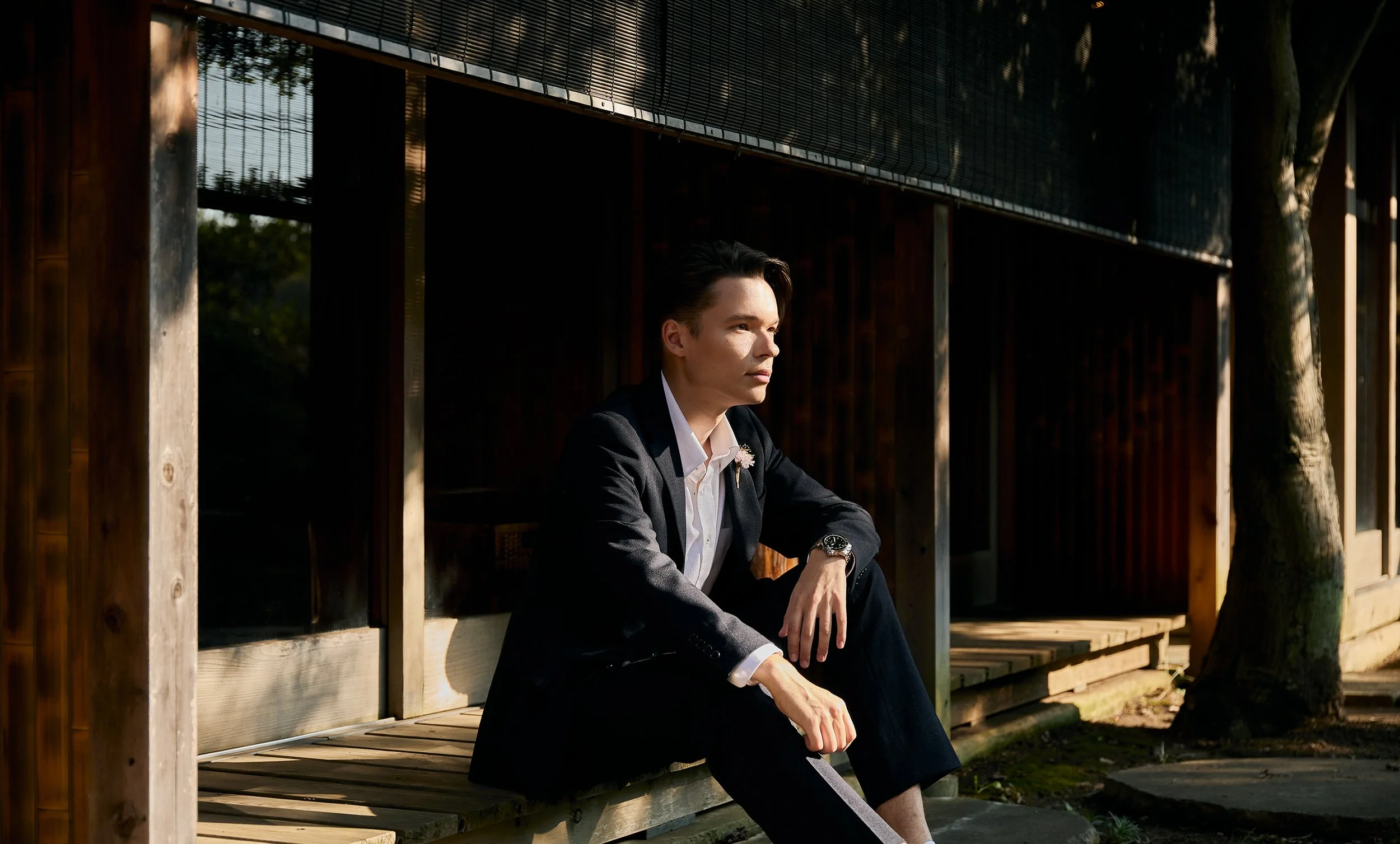 A young man in a suit sits on a wooden bench outside a modern building, gazing thoughtfully into the distance, with sunlight and shadows on his face.
