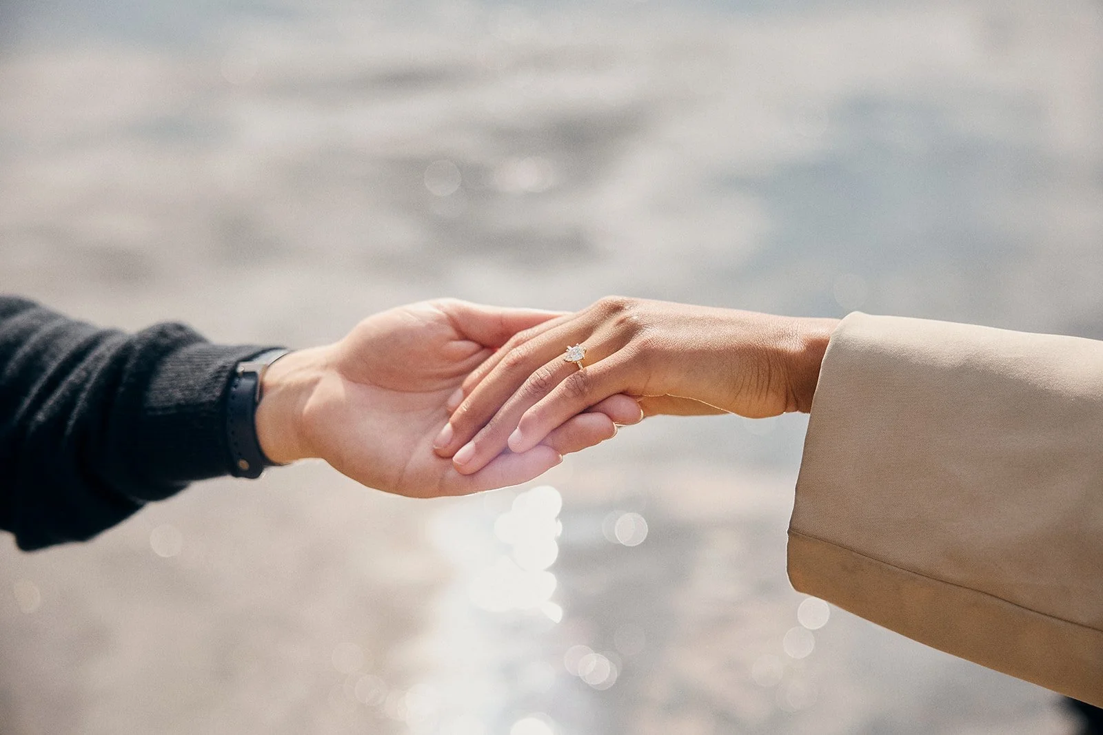 A person is holding another person's hand over water, showing an engagement ring.