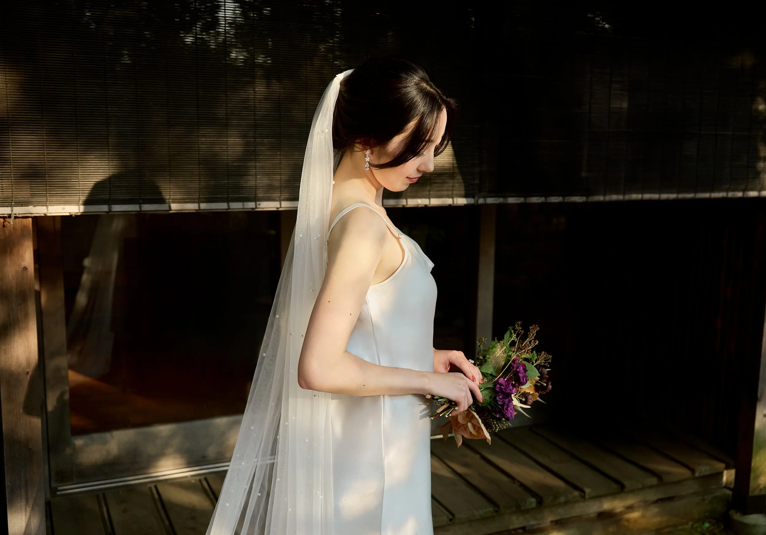 A bride in a white wedding dress and veil holding a bouquet, looking down outdoors during daytime.