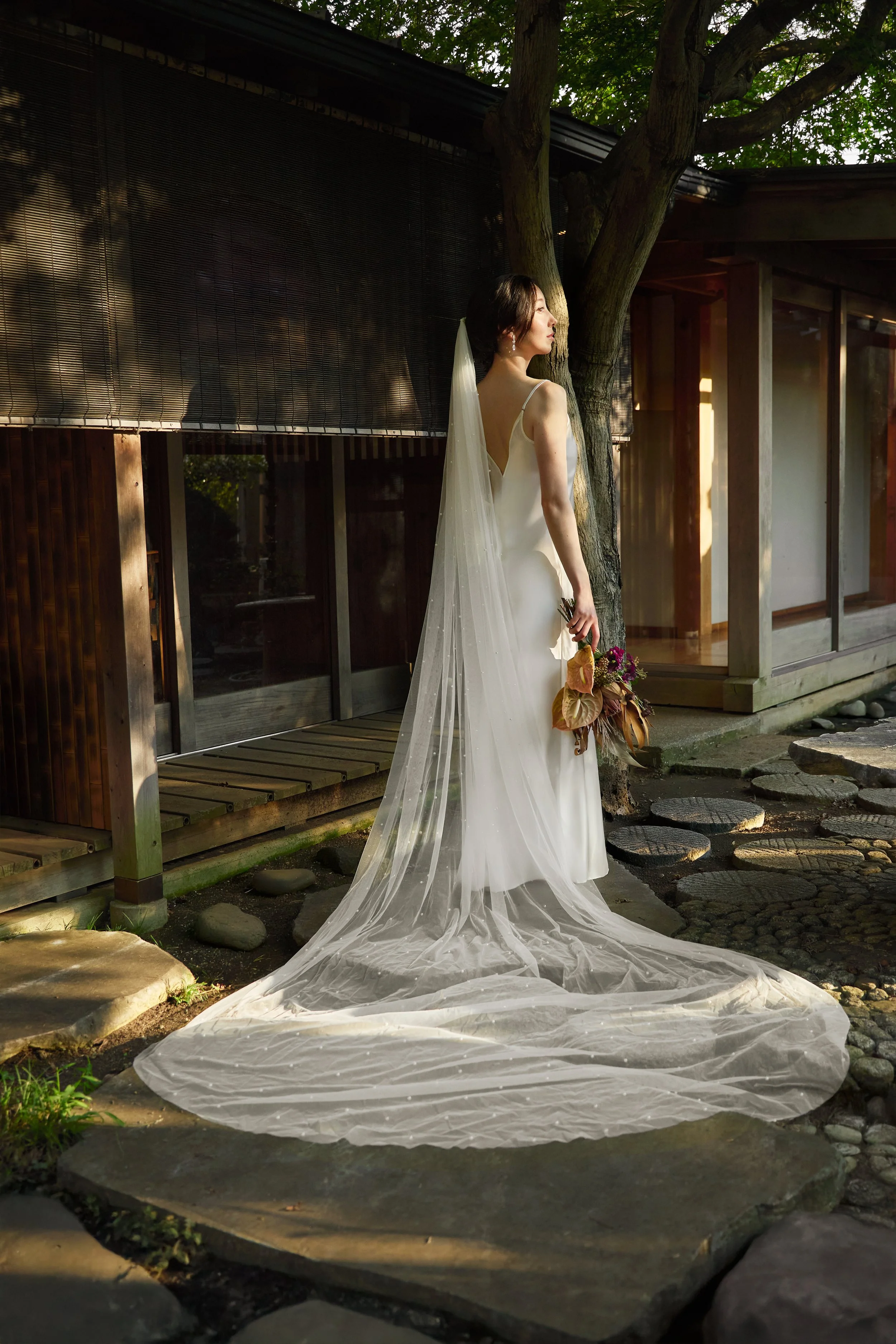 A woman in a white wedding dress with a long veil, holding a bouquet, standing outside near a building and a tree, with sunlight casting shadows.