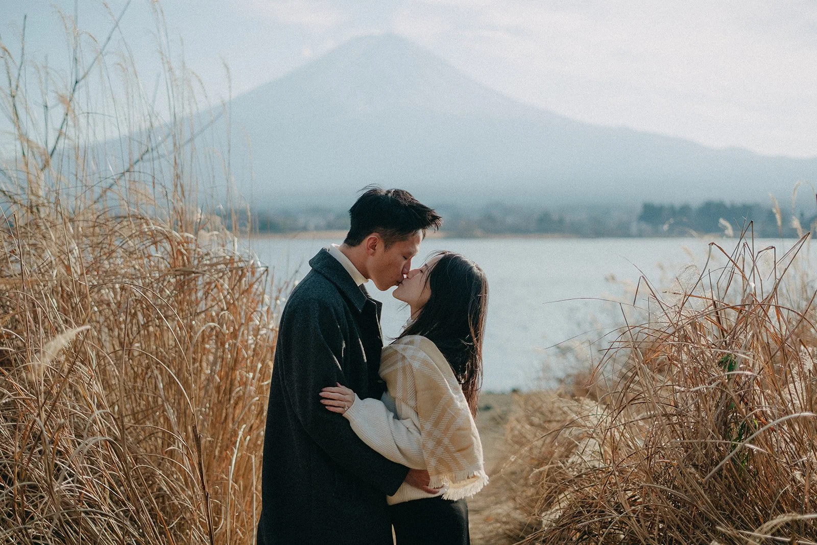 A couple sharing a kiss in a natural outdoor setting near a body of water, with dry tall grasses in the foreground and a mountain in the background.