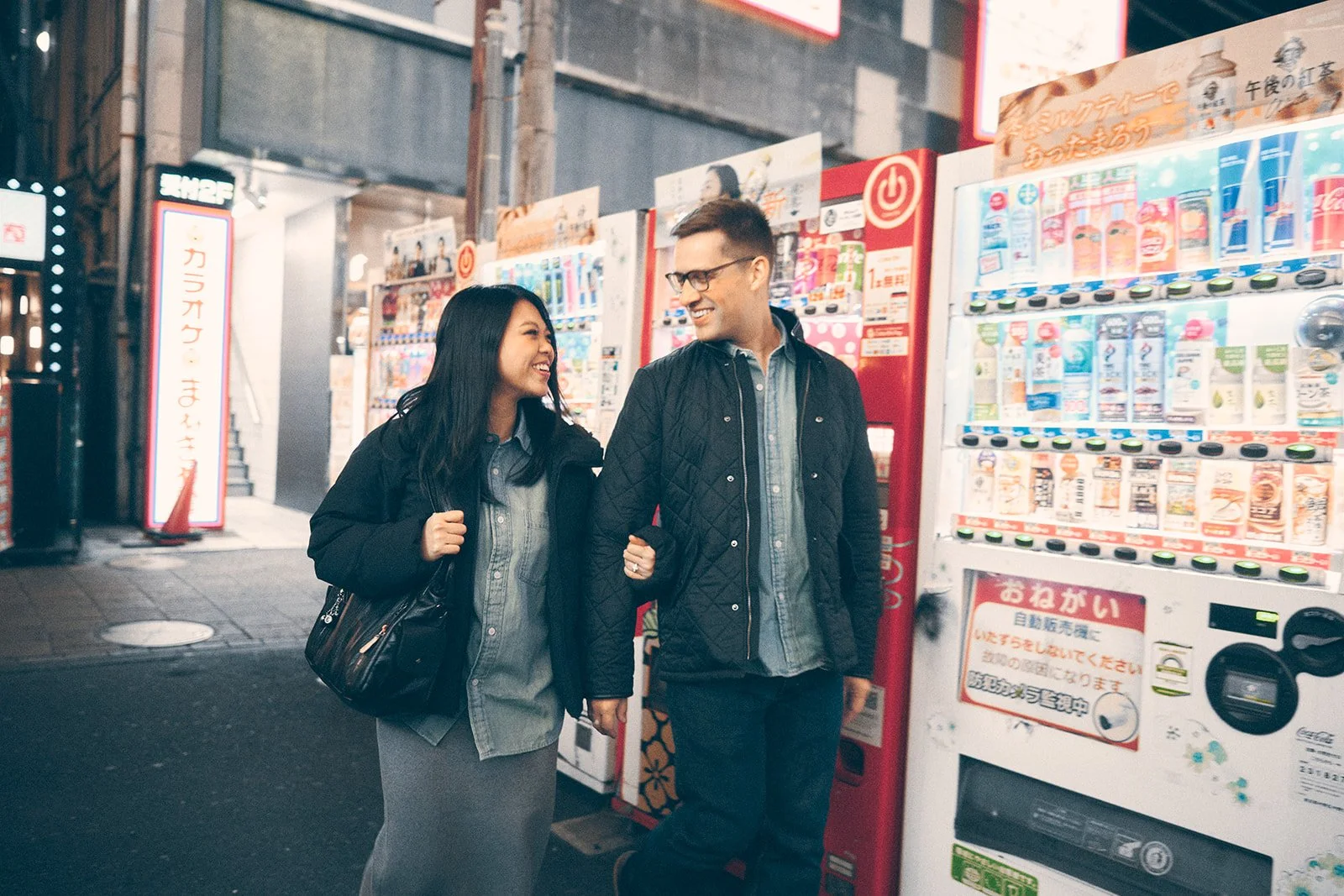 A man and woman smiling and walking together at night near a vending machine on a city street.