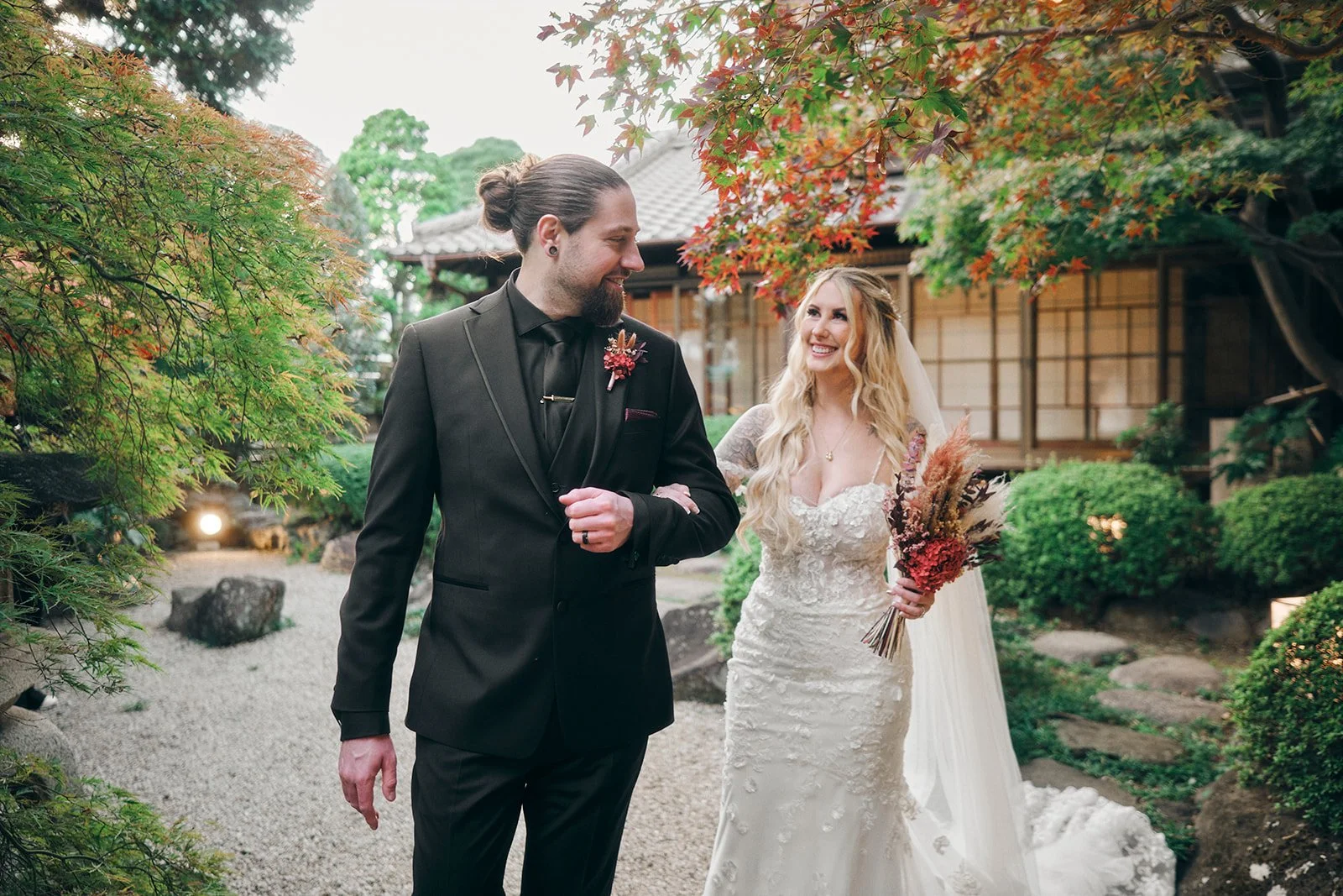 A bride and groom smile at each other outdoors in a garden setting, with trees and a traditional-style building in the background.