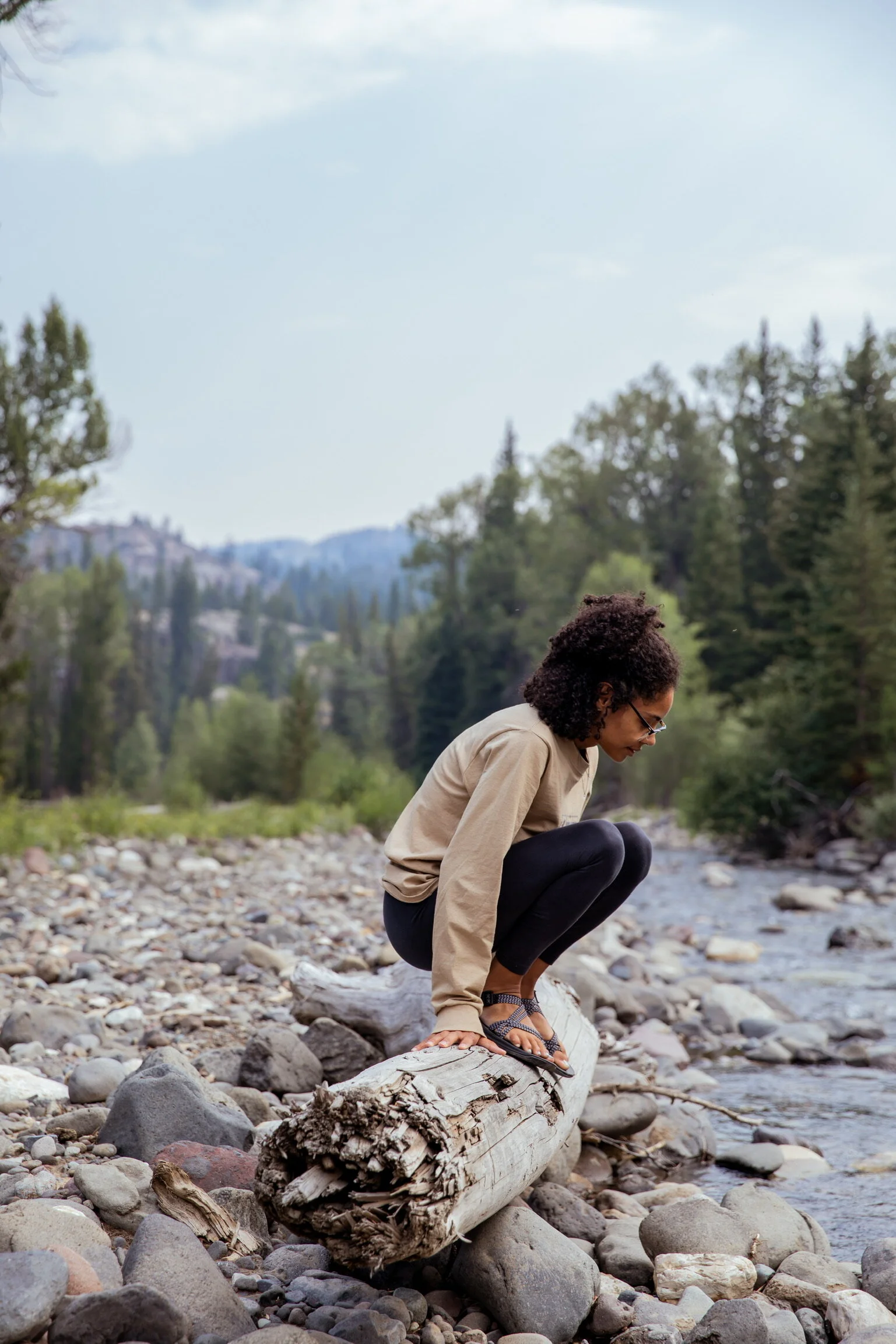 Person crouching on a log by a rocky riverbank, surrounded by forested mountains and trees.