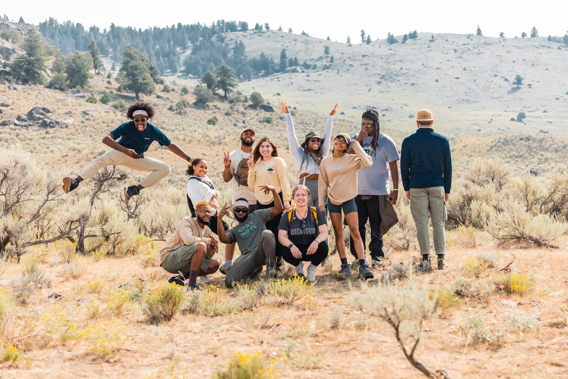 A group of diverse people in outdoor hiking gear posing and playing in a dry, mountainous landscape with sparse bushes and trees.