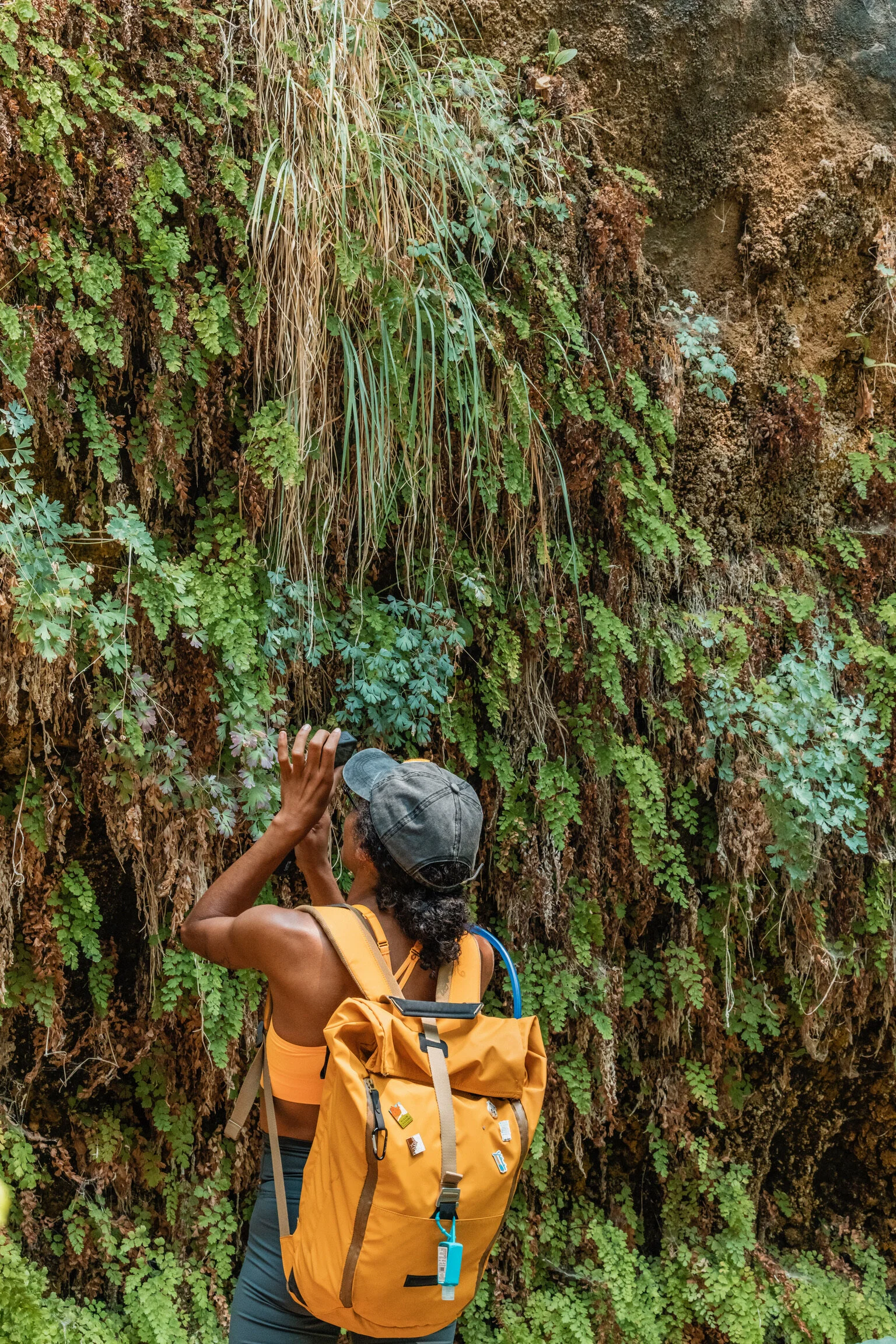 A woman wearing a cap, orange sports bra, and carrying a yellow backpack takes a photo of a moss-covered rock wall with green plants.