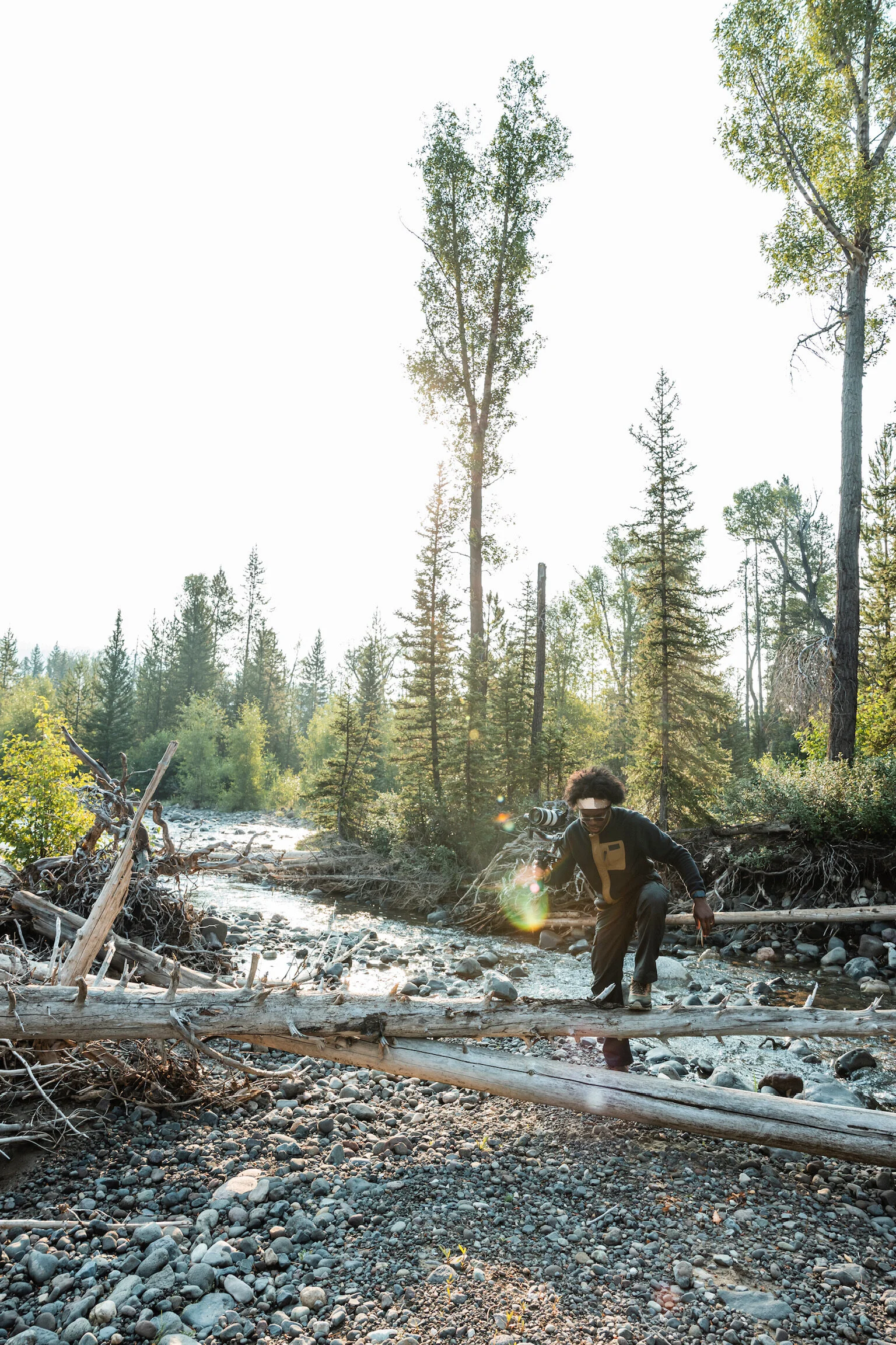 A person with dreadlocks crossing a fallen log over a rocky creek in a forest with tall trees and sunlight streaming through.
