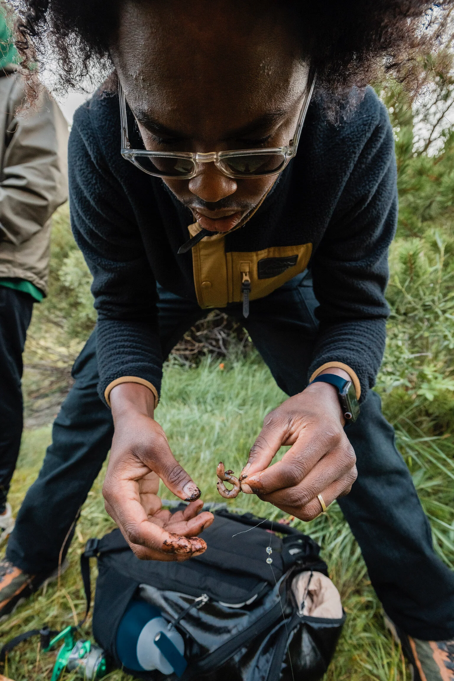A person with curly hair and glasses is crouching outdoors, holding a small insect or caterpillar with hands covered in mud or dirt. There is greenery and another person partially visible in the background, along with a black backpack on the ground.