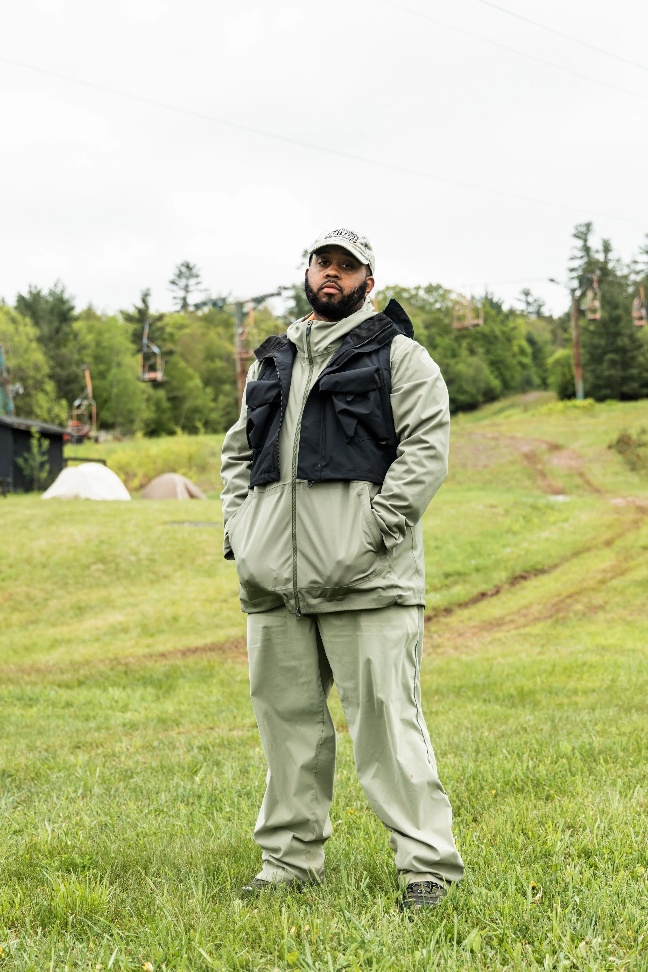 A man standing outdoors on a grassy area with tents and trees in the background, dressed in outdoor gear including a beige jacket, beige pants, a black vest, and a cap.
