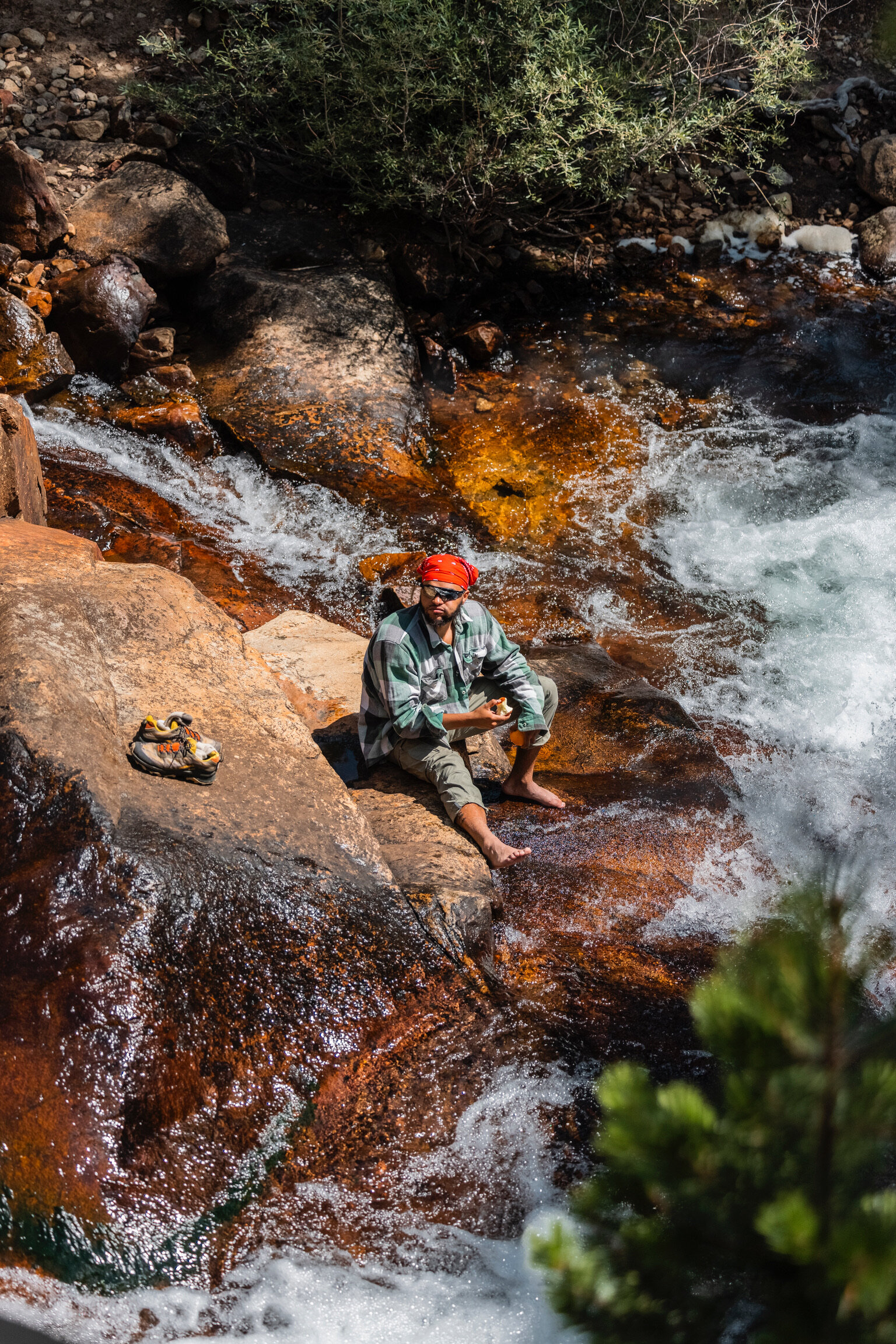 A man with a red bandana and sunglasses sitting on a large rock by a rushing river, eating a snack, with hiking shoes placed nearby. Green foliage is visible in the background.