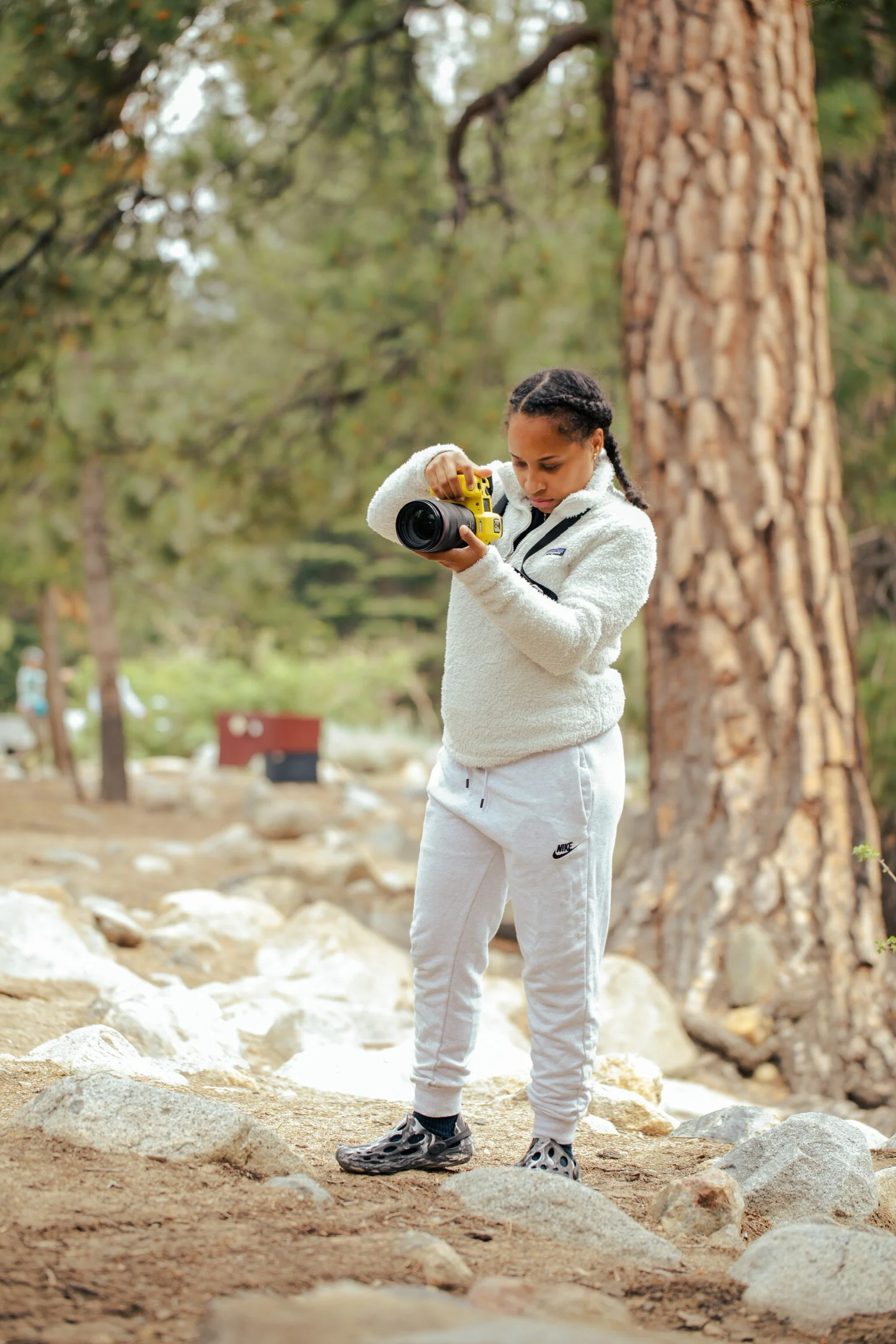 A young girl wearing a fluffy white jacket, light gray Nike sweatpants, and black Crocs is standing outdoors on rocky terrain, looking at a camera she is holding. She is in a forested area with tall trees and green foliage.