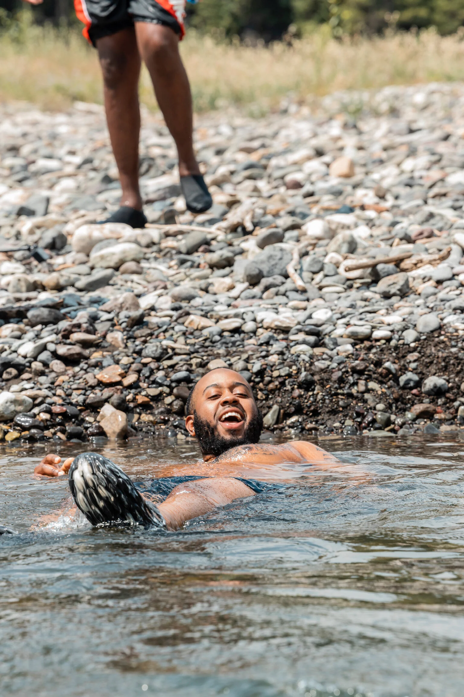 A man with a beard is smiling and floating in a body of water near a rocky shoreline. Another person, wearing black shoes and shorts, is walking on the rocks in the background.