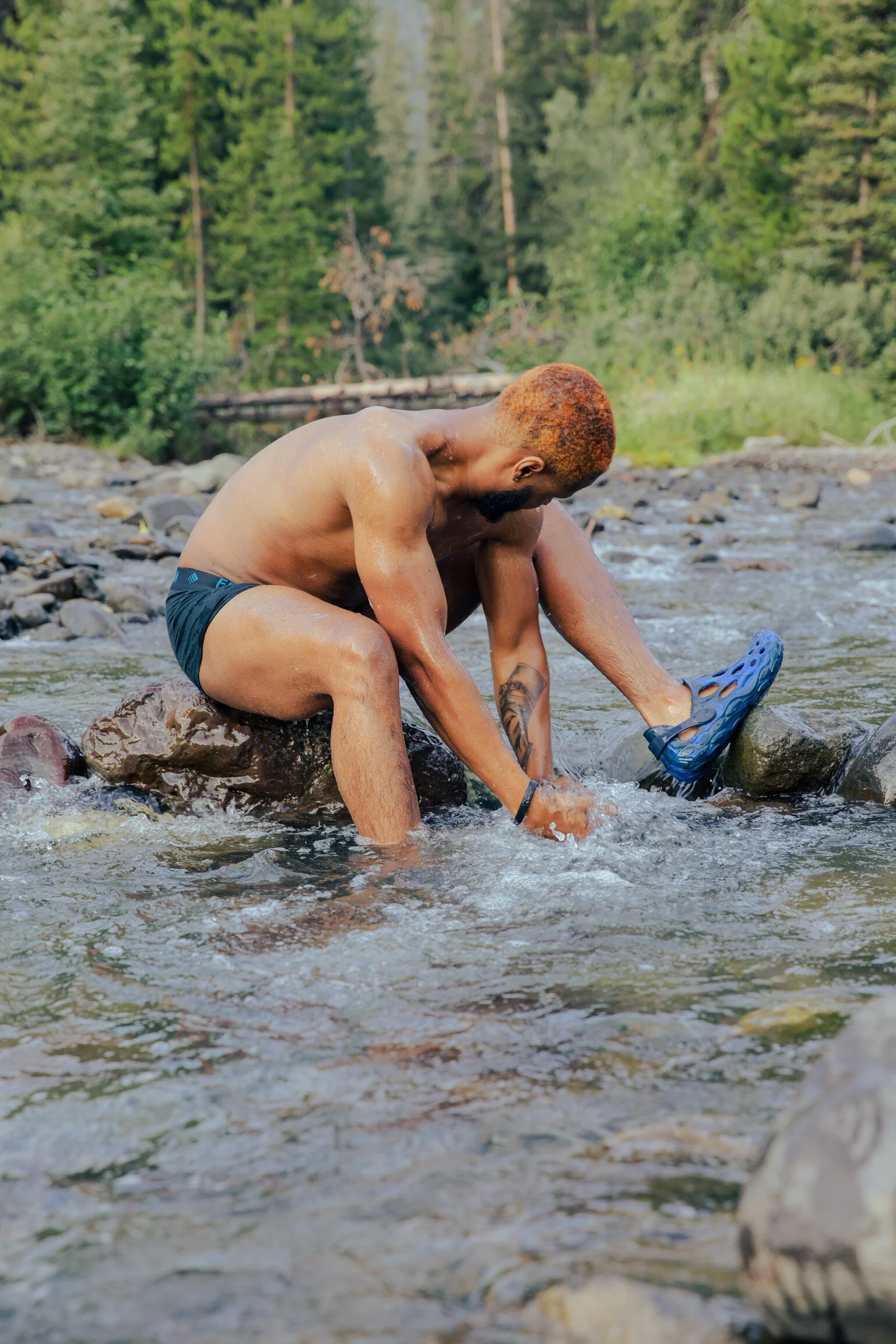 A shirtless man with orange hair and tattoos on his arm sitting on a rock in a shallow creek, putting on or taking off a blue sandal, surrounded by trees and rocks.