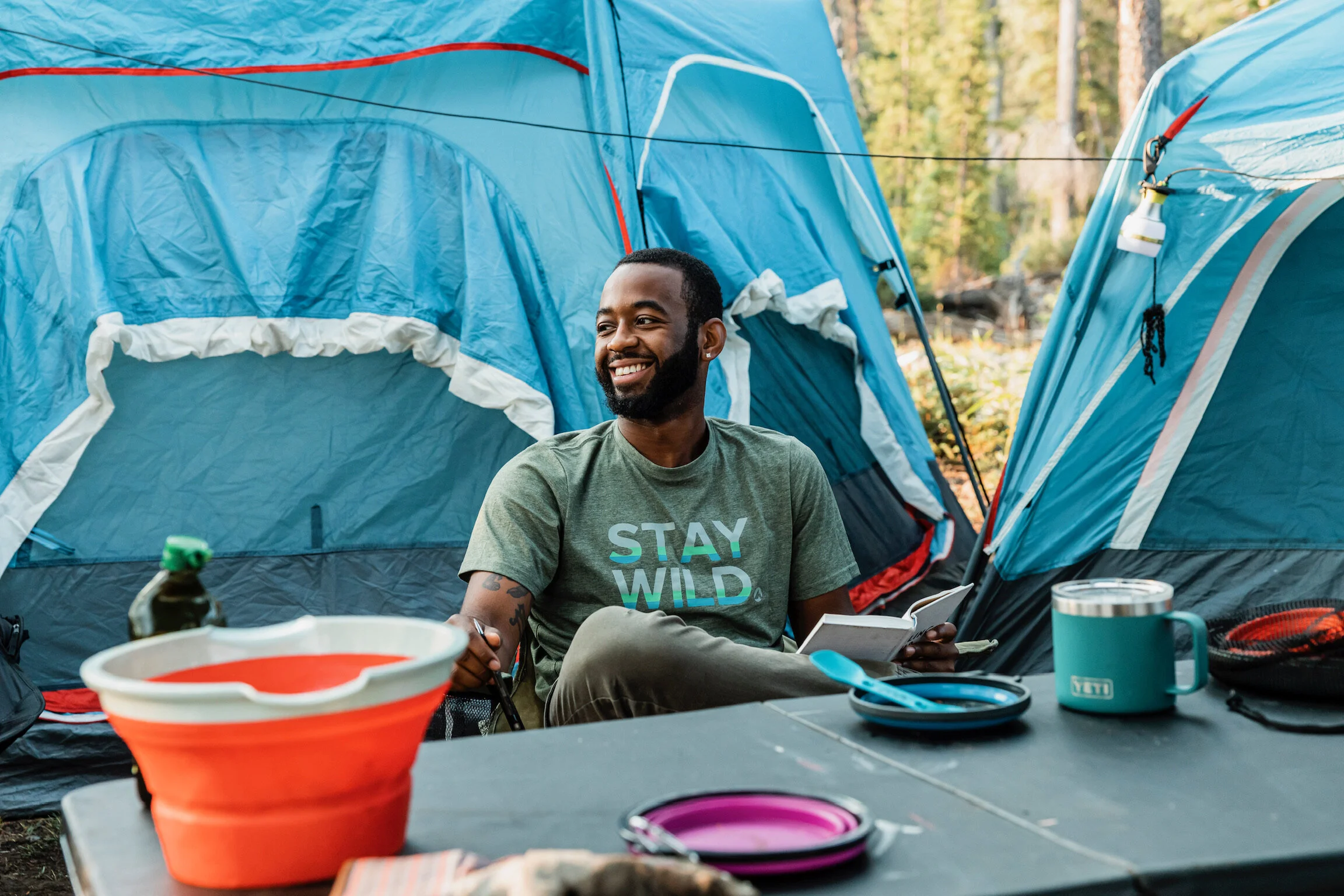 A man sitting at a campsite table outdoors with blue camping tents in the background, smiling and looking to the side, wearing a t-shirt that says 'STAY WILD' with camping gear on the table.