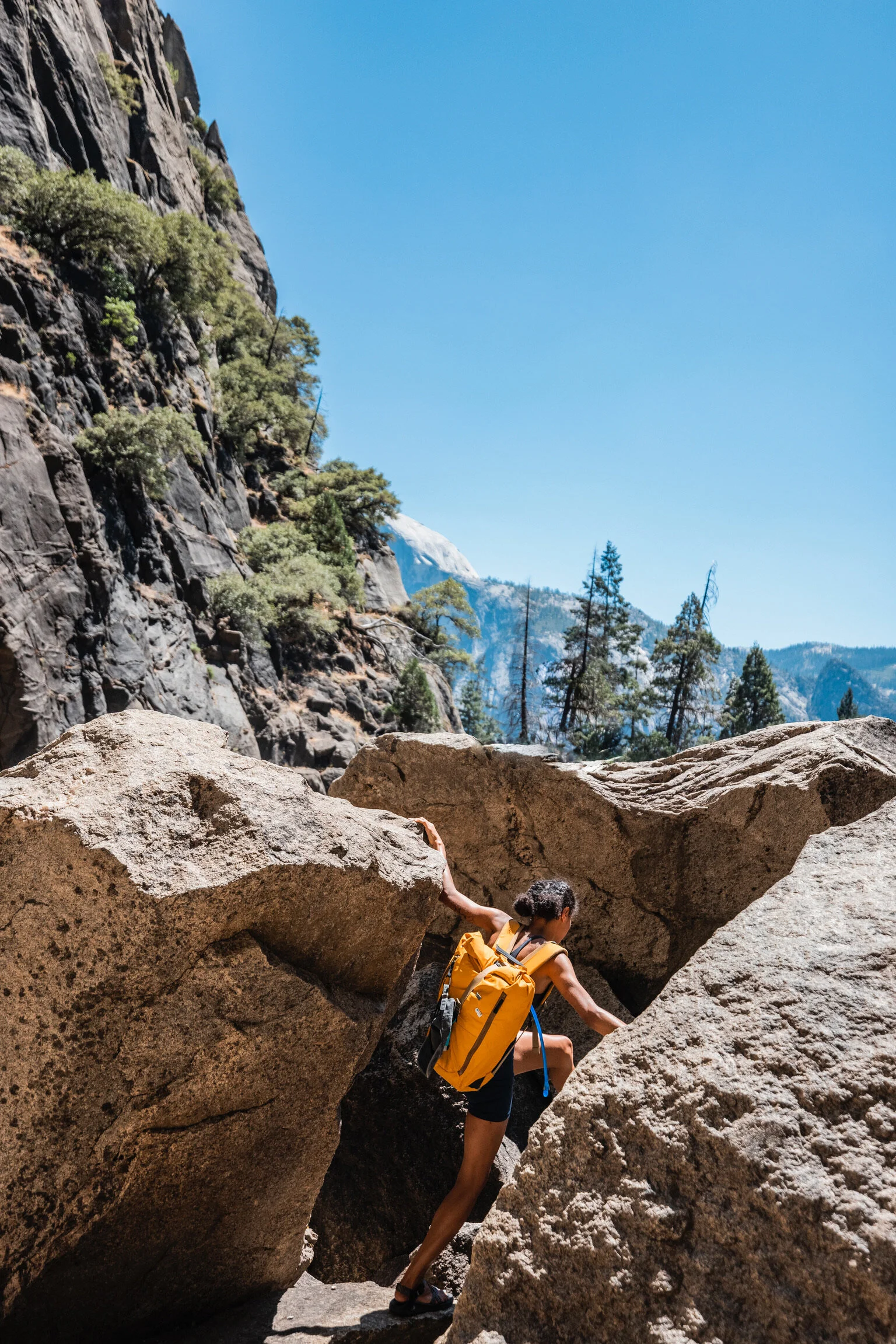 A woman with a yellow backpack hiking through a rocky canyon with large boulders and trees, mountains in the background under a clear blue sky.