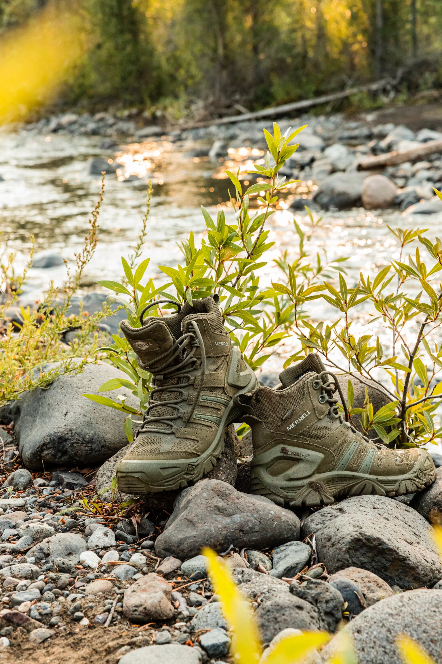 A pair of green hiking boots placed among rocks by a river with trees and sunlight in the background.