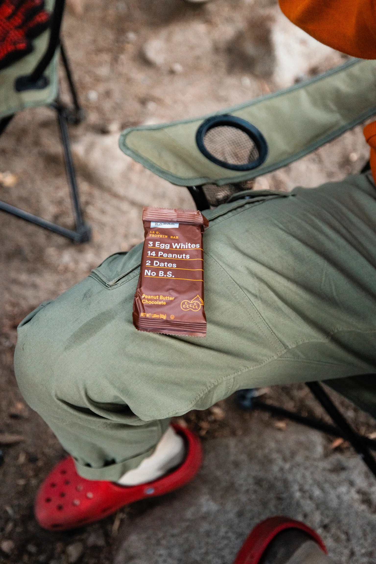 A person seated outdoors wearing green pants and red Crocs, with a protein bar resting on their leg. The background shows dirt and a camping chair.