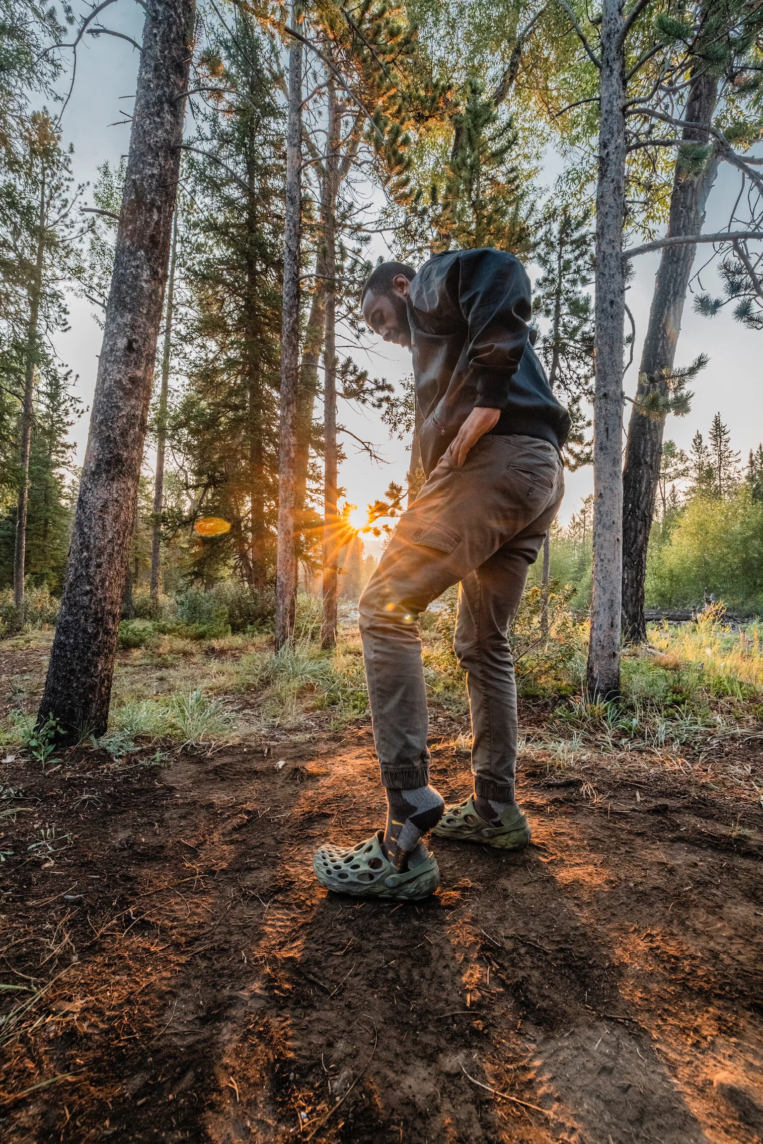 A man standing on a dirt trail in a forest during sunset, looking down, wearing tan cargo pants, a black hoodie, and sandals, with trees and sunlight in the background.