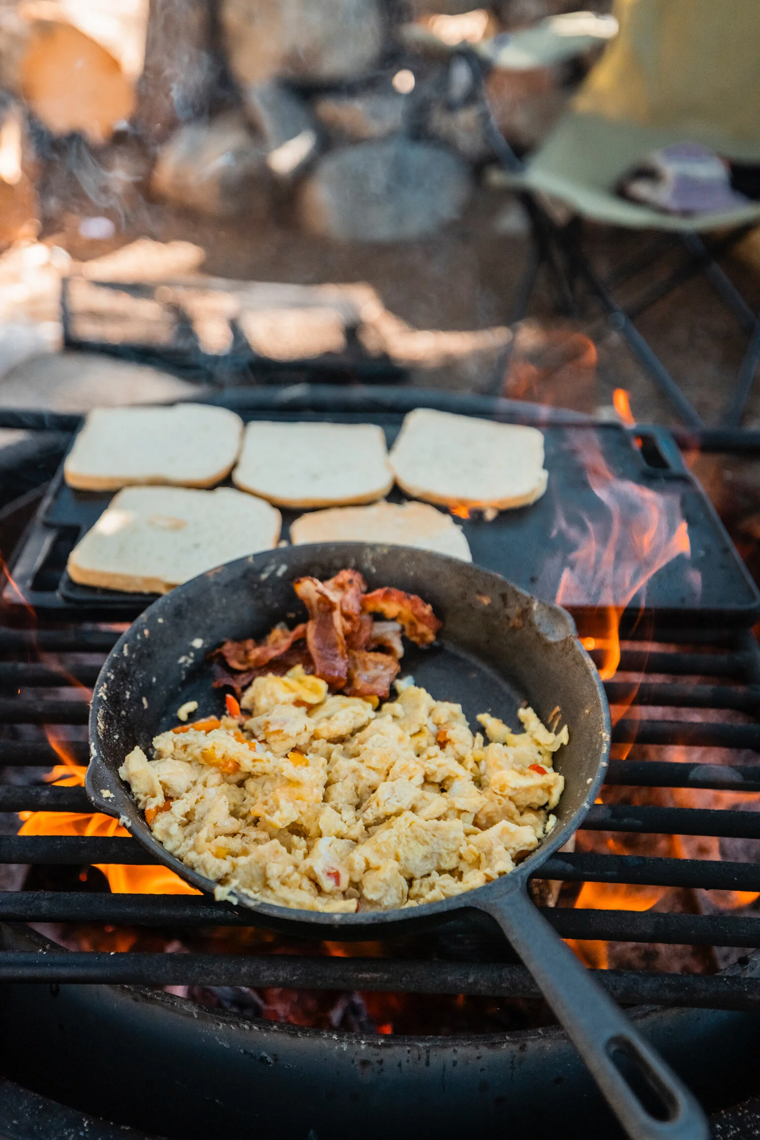 Cooking breakfast over an open campfire with scrambled eggs and bacon in a frying pan, slices of bread toasting on a grill, outdoor camping setup with chairs in the background.