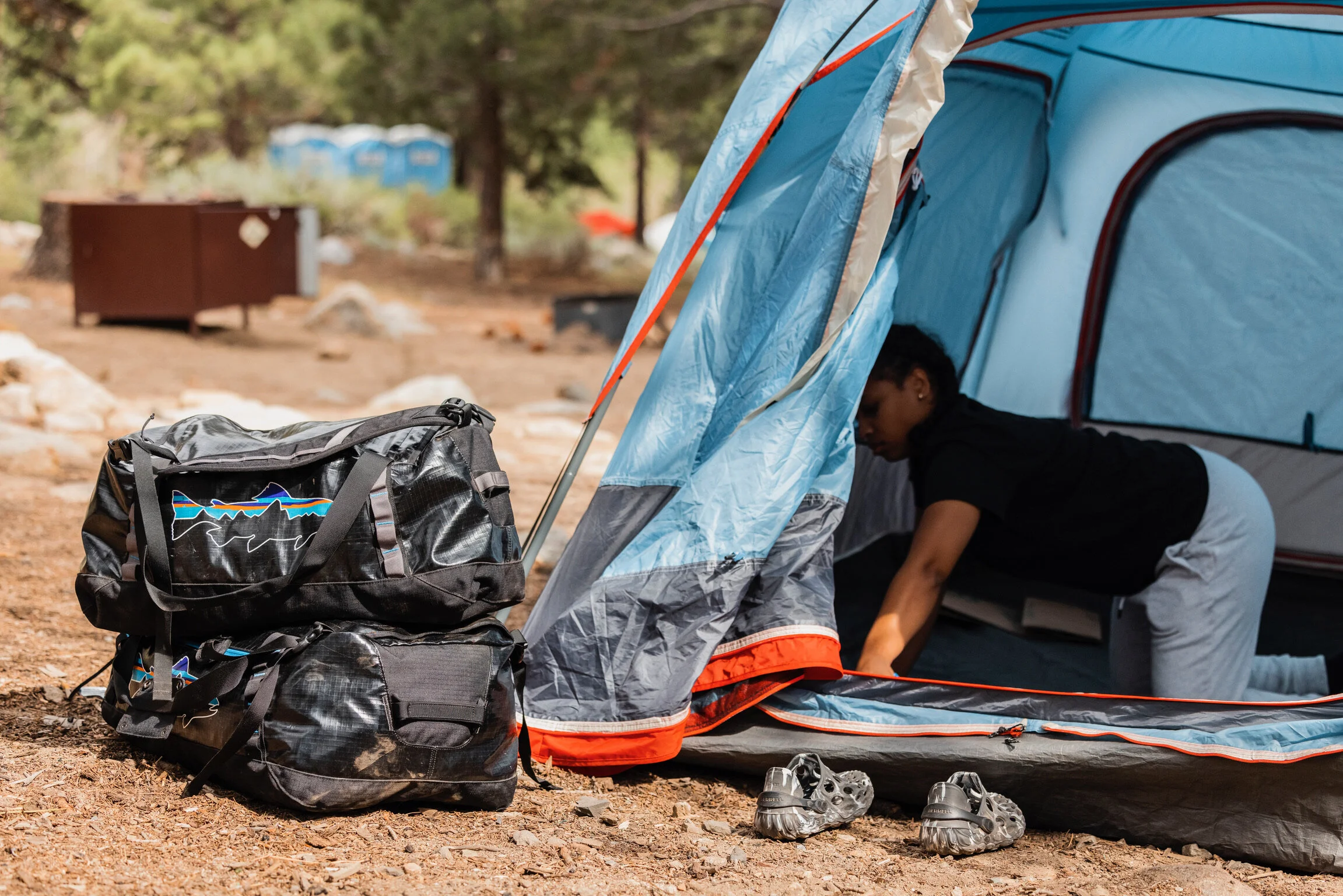 A person setting up a blue camping tent outdoors with two backpacks and a pair of shoes nearby.