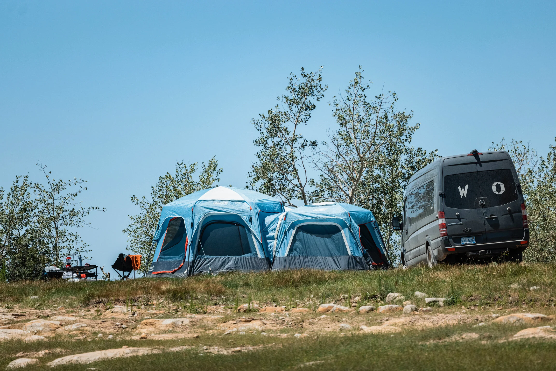 Camping scene with a blue tent, a black van with 'W O' written on the back window, a camp chair, and some trees under a clear blue sky.
