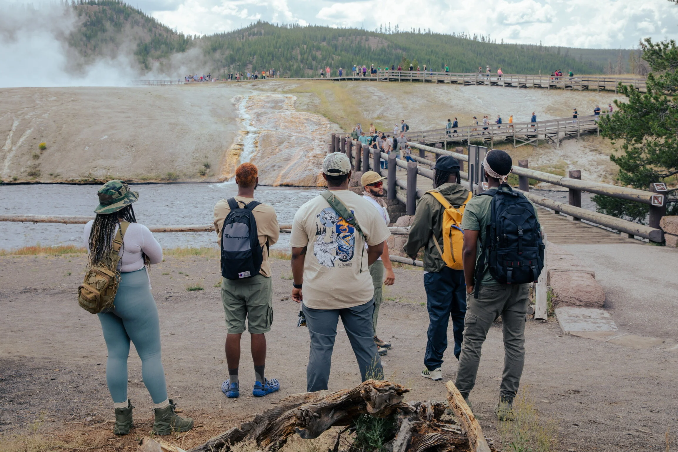 Group of people observing a geyser at a geothermal area, with steam rising in the background and a wooden walkway leading toward the geyser.