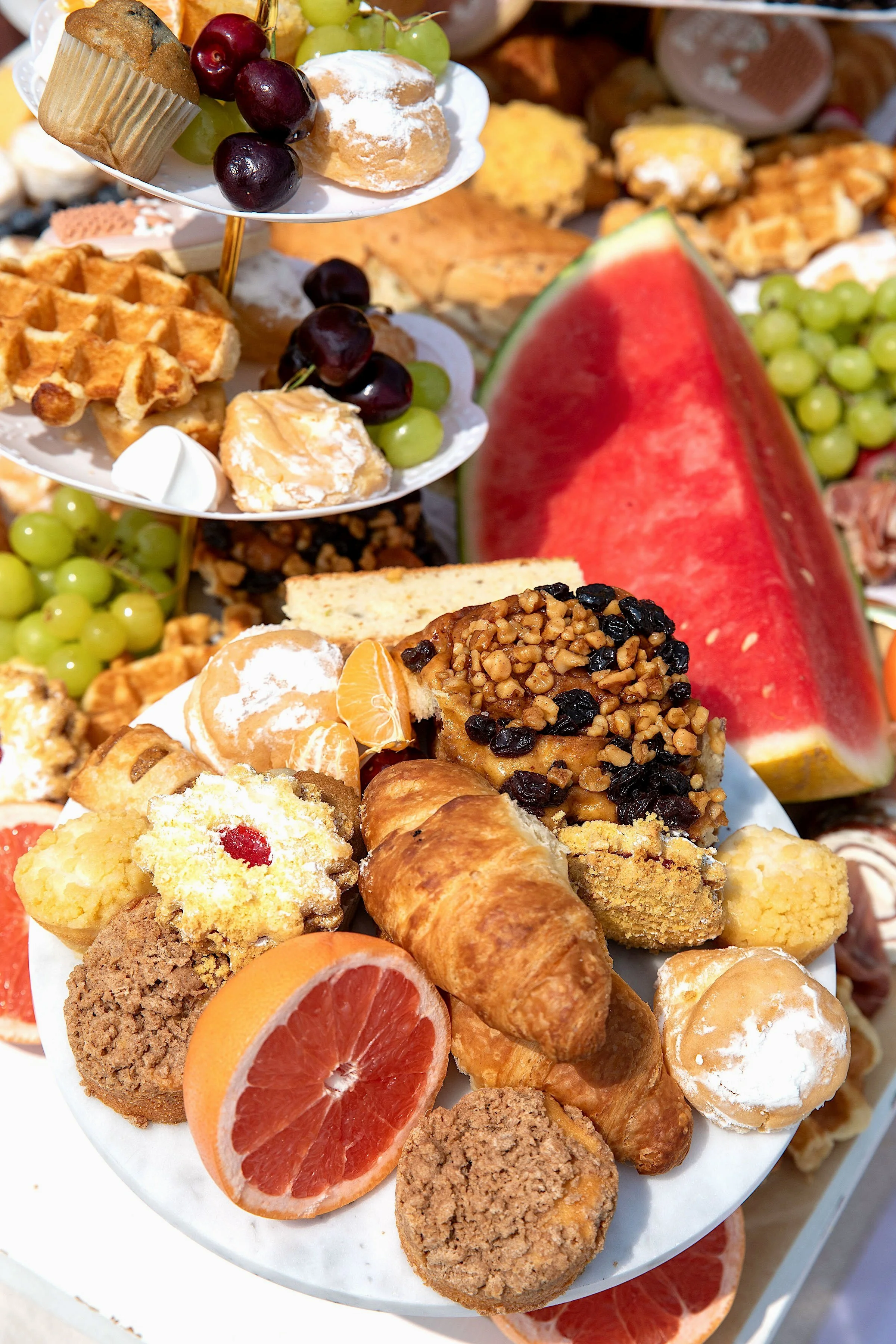 Assorted desserts and fresh fruit on a platter, including croissants, cookies, slices of watermelon and grapefruit, grapes, and small pastries.