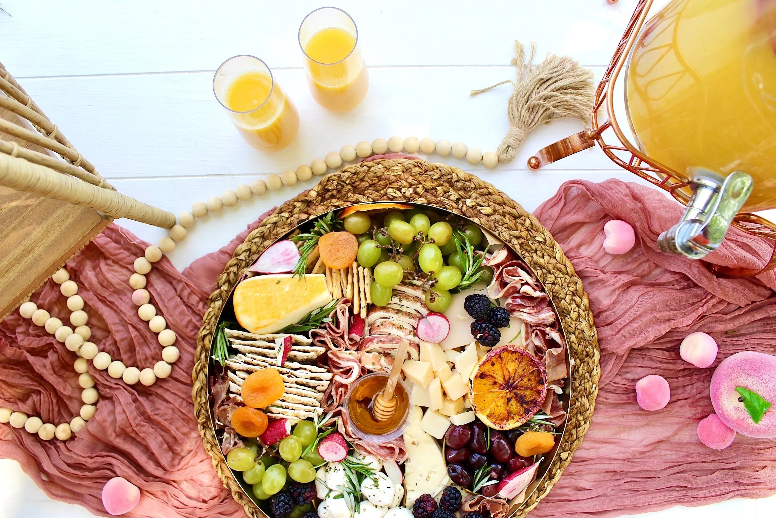 Charcuterie board with cheese, grapes, meats, and dried fruits, along with two glasses of orange juice, on a white table with pink and beige decorations.