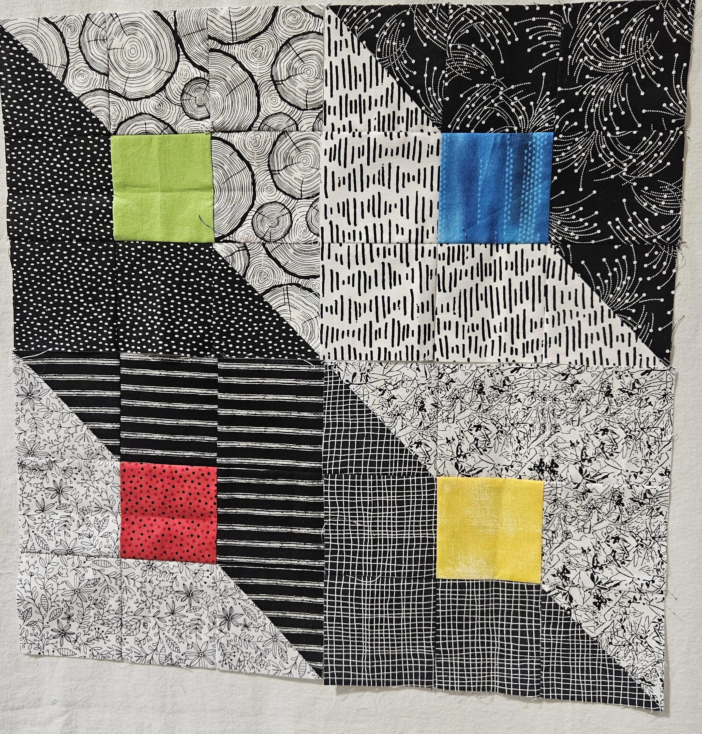 Four squares of bright colours (blue, green, red and yellow) surrounded by a mostly black fabric with white accents, and mostly white fabric with black accents, that are cut diagonally