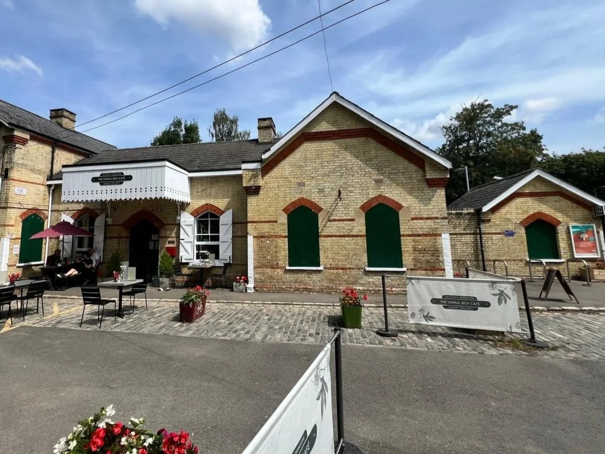 Exterior view of The Signal Box Cafe, a brick building with green shutters and white trim, outdoor seating with tables, chairs, and umbrellas, flower pots, and a cobblestone sidewalk under a partly cloudy sky.