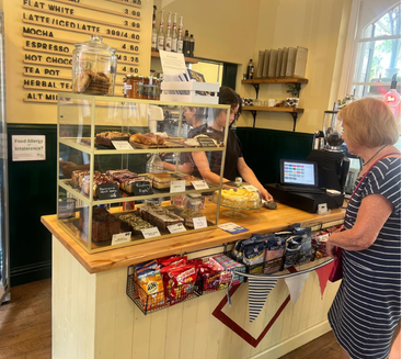 A woman in a striped shirt stands at a coffee shop counter with a glass display case filled with assorted baked goods, including brownies, cookies, and bars. Behind the counter, a barista prepares drinks. A menu board displays beverage options and prices. Shelves with coffee supplies are visible in the background, and a customer with short blond hair waits at the counter.