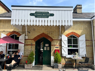 Exterior of The Signal Box Cafe, a brick building with a white awning and green sign. There are open white shutters, potted plants, and outdoor seating with several people sitting and relaxing on a sunny day.