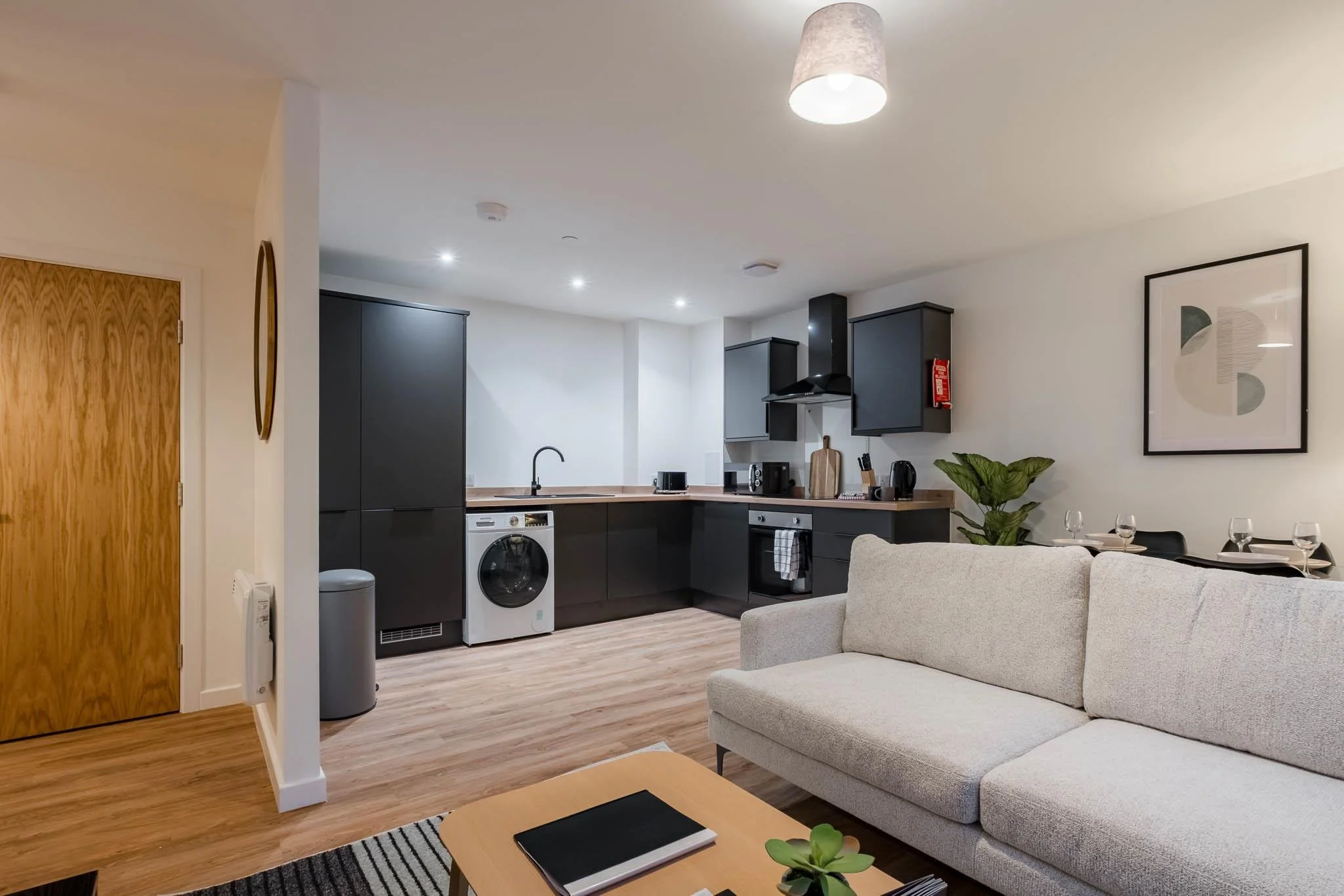 Open-concept living room and kitchen with beige sofa, wooden coffee table, dining table with wine glasses, modern black and white kitchen cabinets, appliances, and wall art.