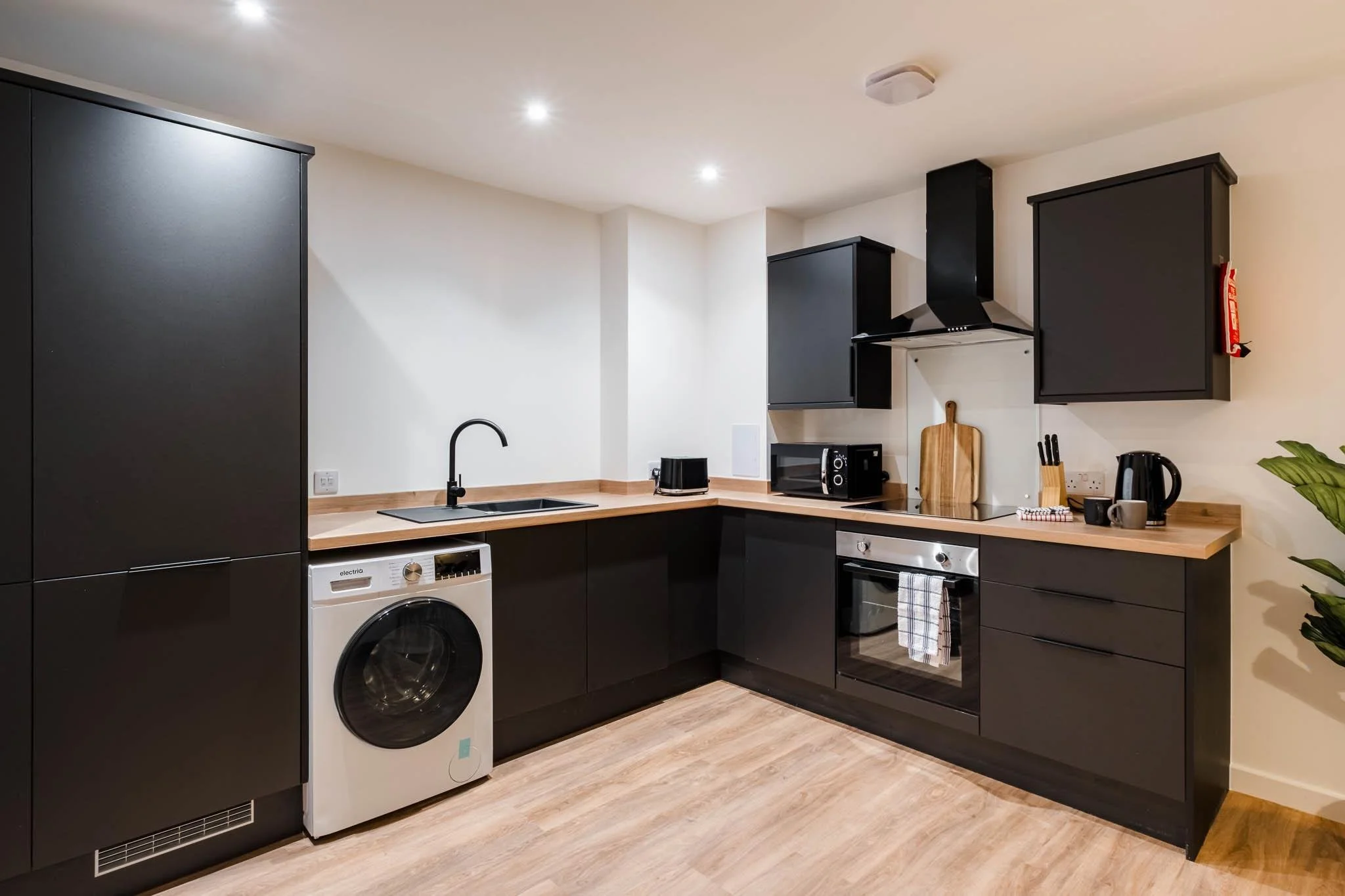Modern kitchen with black cabinets, wooden countertop, washing machine, microwave, toaster, kettle, and various small appliances.